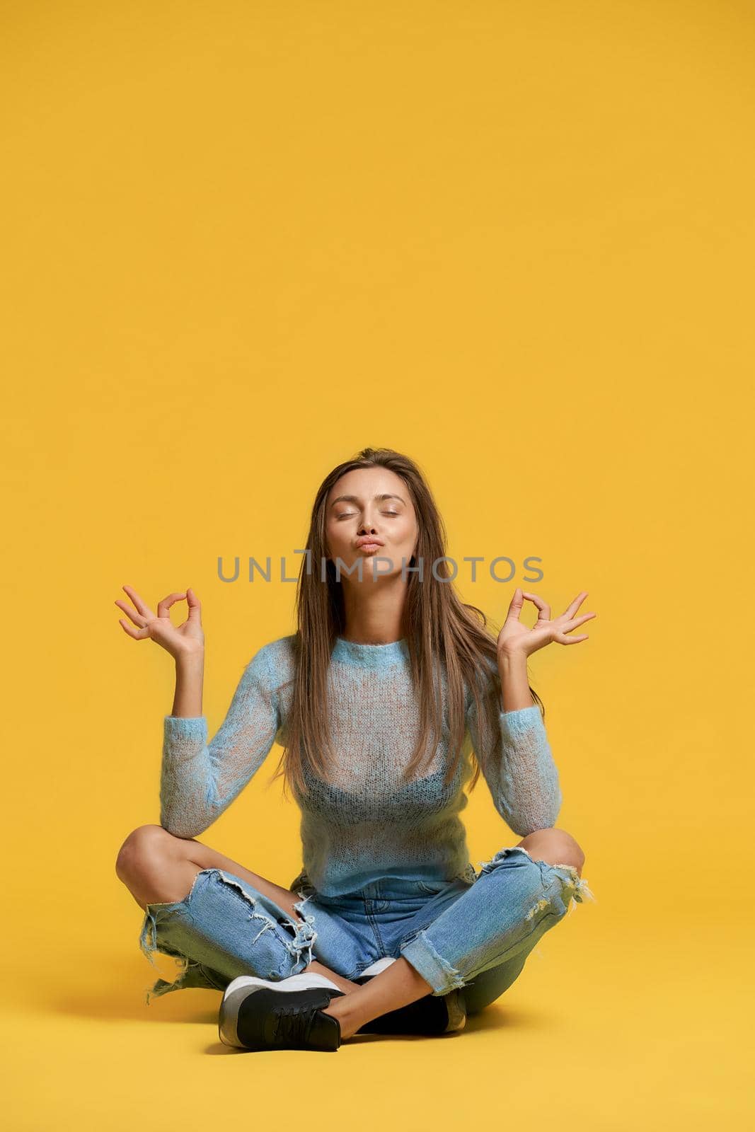 Front view of girl showing peace using lotus pose. Stylish pretty brunette woman sitting on floor using fingers to show balance, making duck lips on yellow background. Concept of fun, meditation.