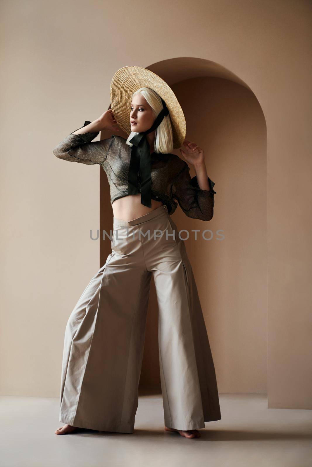 Attractive blonde woman wearing tied up blouse and wide beige trousers holding hand on her trendy straw and posing on camera. Young girl with bare torso and feet standing near arch in the wall