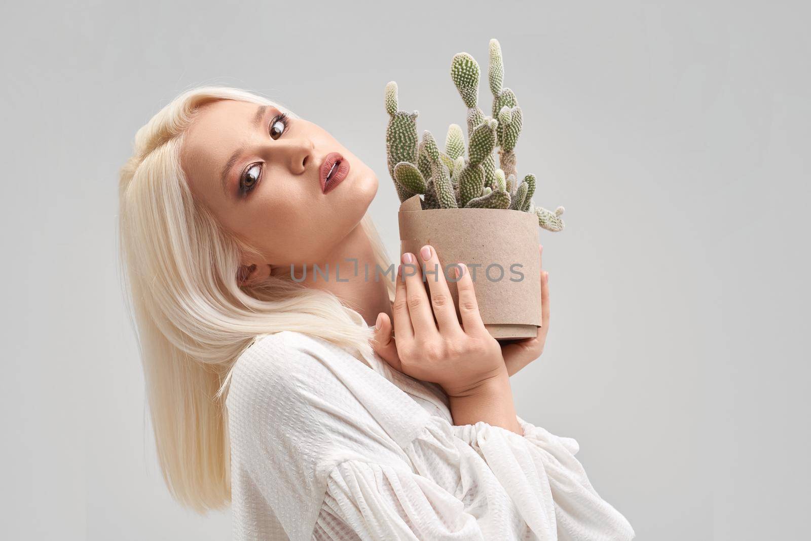Portrait of beautiful blonde girl with make up and hairstyle wearing white blouse, looking at camera and holding pot with green cactus. Pretty young woman buying plant for house