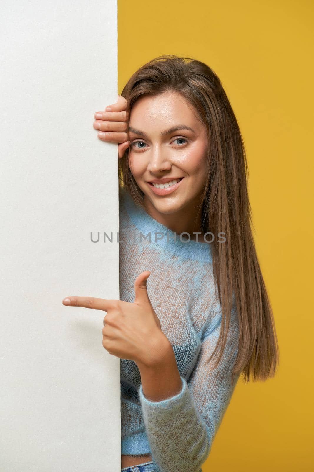 Front view of smiling pretty girl with long brown hair peeping out of white poster, pointing at poster looking at camera wearing jeans.