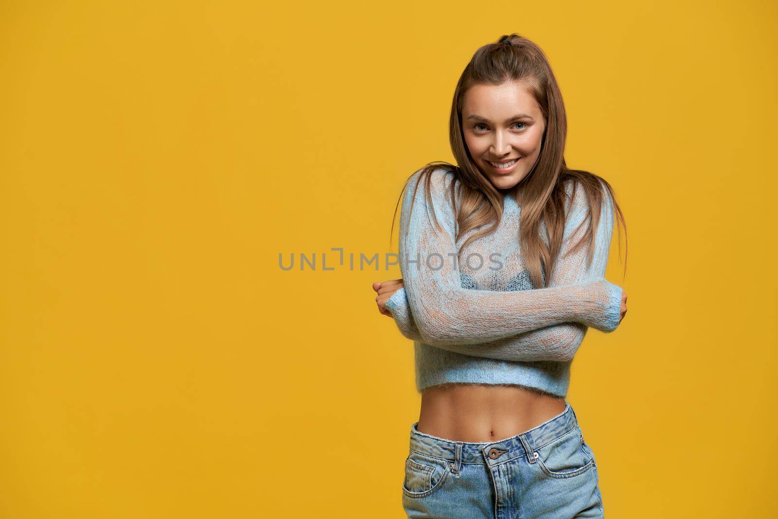 Front view of pretty smiling girl with long brown hair hugging herself. Young lady wearing short blue top with long sleeves on isolated yellow background at studio. Concept of beauty, fashion, style