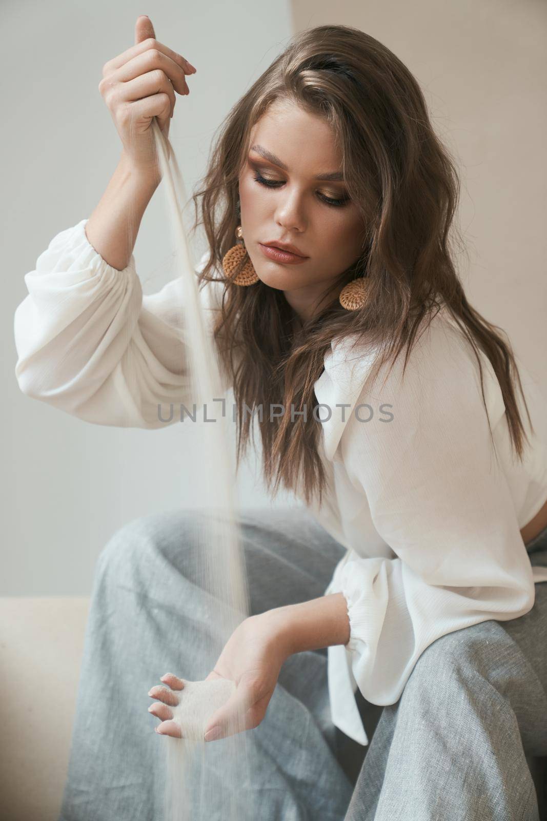 Attractive professional model with trendy make up and hairstyle looks very stylish in white blouse and grey trousers while pouring sea sand from one hand to another. Fashion looks. Modeling concept