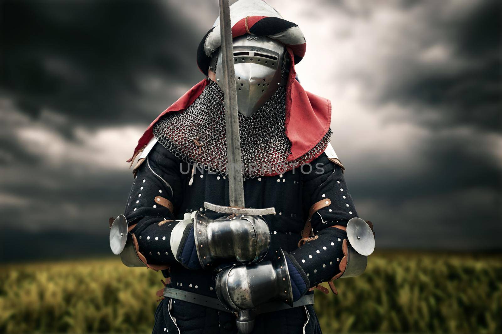 Front view of medieval knight in iron armor and headband posing and holding big sword. Portrait of brave man standing in field while sky is gray and cloudy. Concept of warrior, middle ages.
