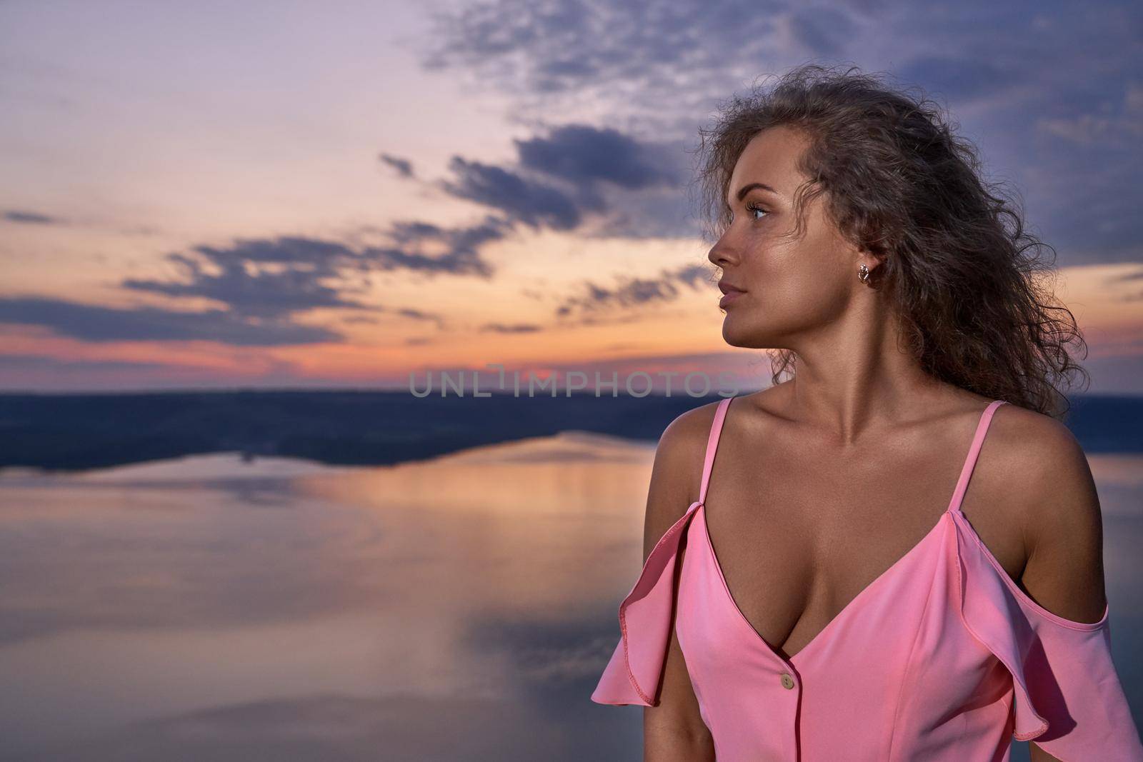 Stylish, seductive lady with curly hair on background of lake and sky in evening. Attractive, romantic girl wearing in pink dress with bare shoulders and dekollete looking away.