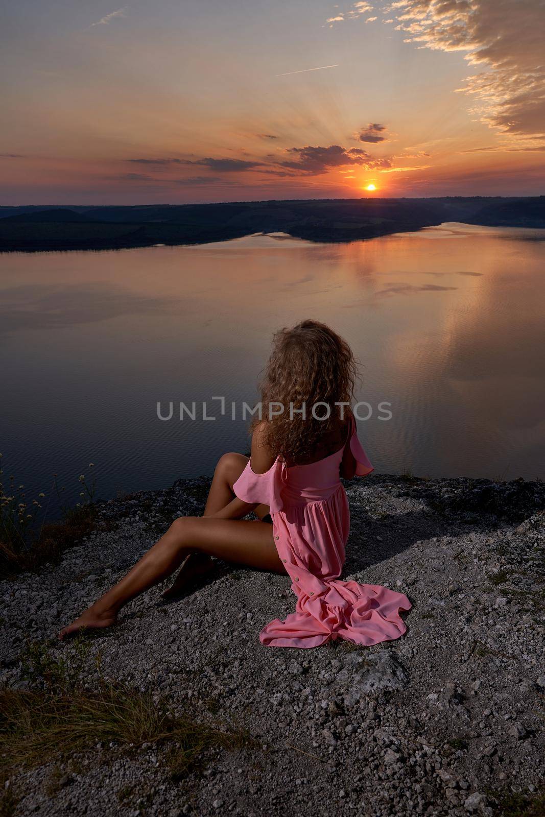 Passionate girl in pink light silk dress sitting on shore of lake on background of sunset. Seductive model with bare shoulders and feet looking at water, admiring sundown.