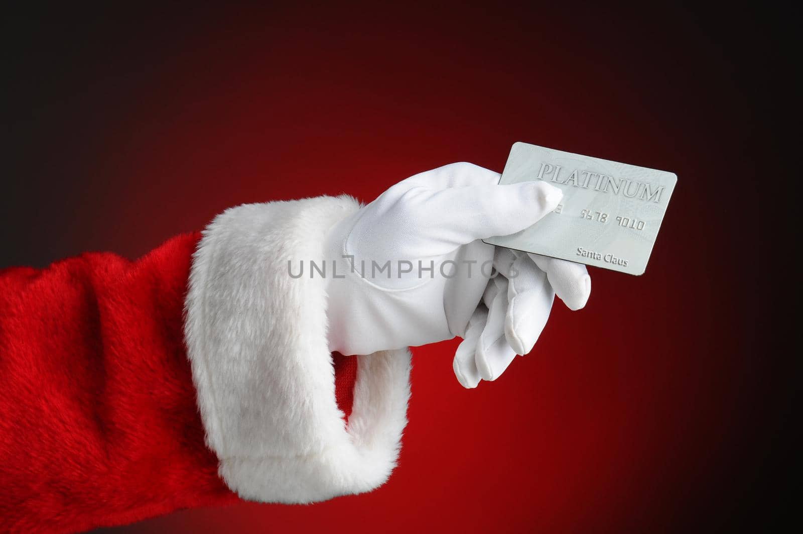 Closeup of Santa Claus hand holding a Platinum Credit Card. Horizontal format over a light to dark red background.