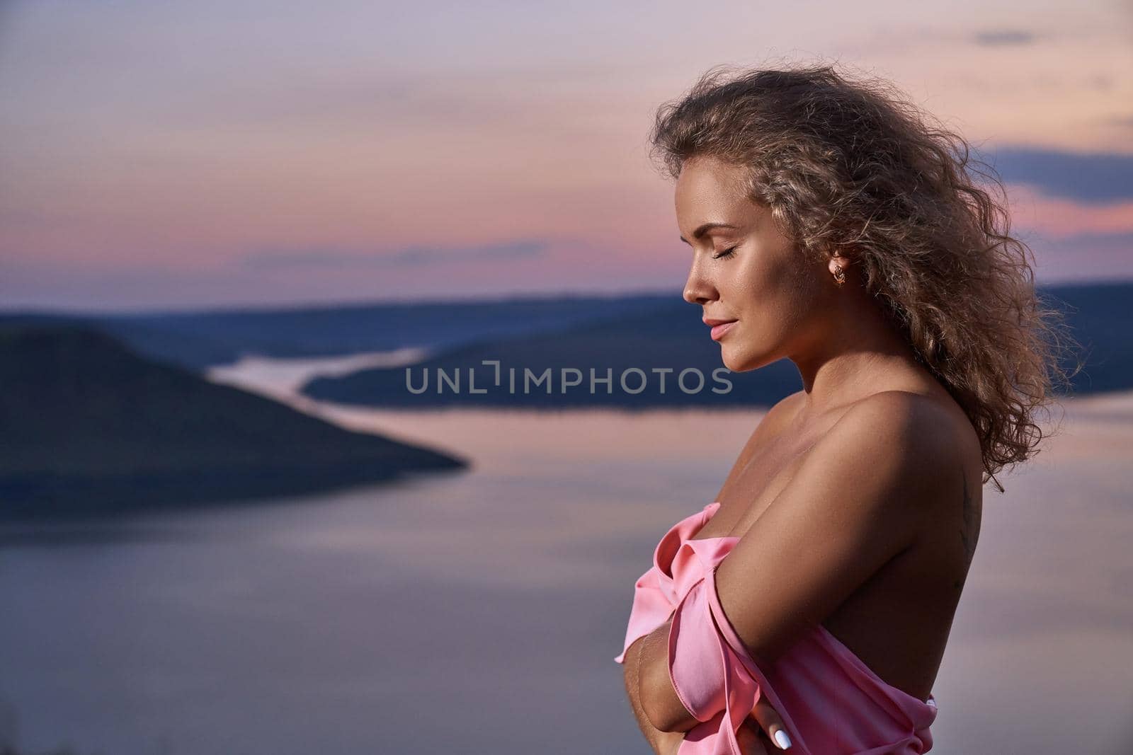 Romantic, charming girl on background of evening lake. Beautiful model with long curly hair with closing eyes wearing in pink dress on background of landscape of sunset.