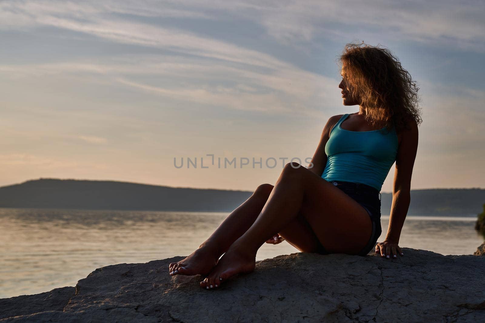 Sexy, beautiful girl with bare feet sitting on stones at shore of lake on sunset. Lady with curly hair wearing in jeans shorts looking at water, admiring landscape.