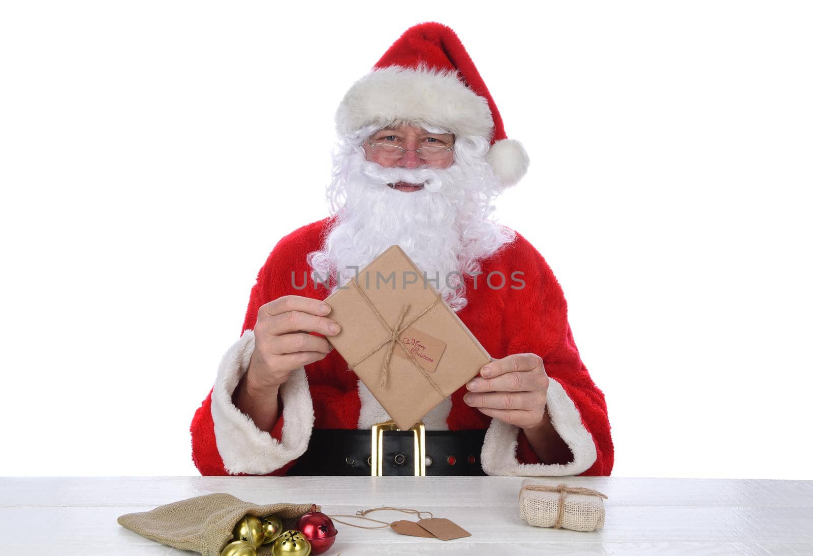 Santa Claus sitting a a rustic white wood table holding a plain paper wrapped Christmas Present, isolated on white.