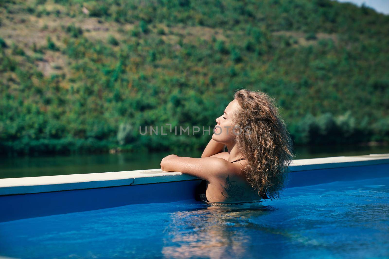 Young seductive girl with blonde long curly hair relaxing in pool of spa resort on nature. Attractive, sexy model in blue water leaning on edge of pool with closed eyes.