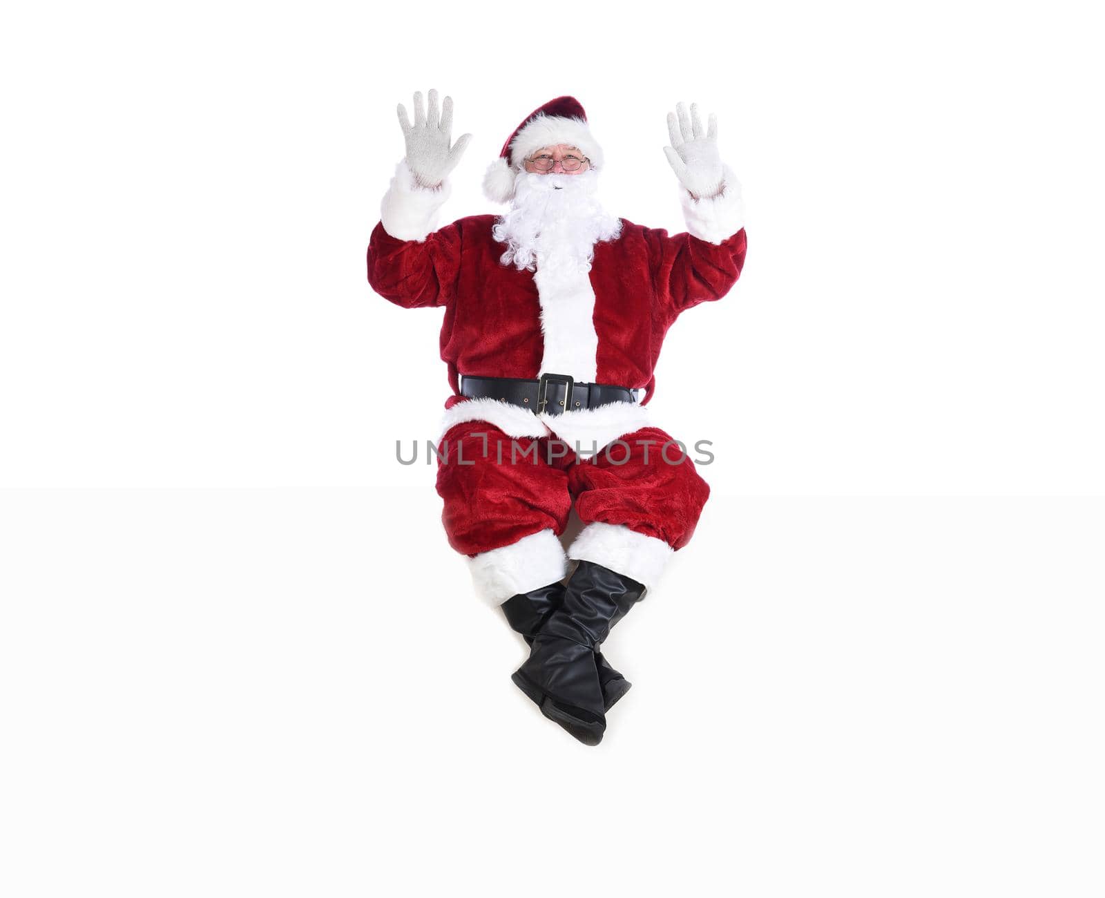 Senior man in traditional Santa Claus Suit sitting on a white wall with both hands in the air. by sCukrov