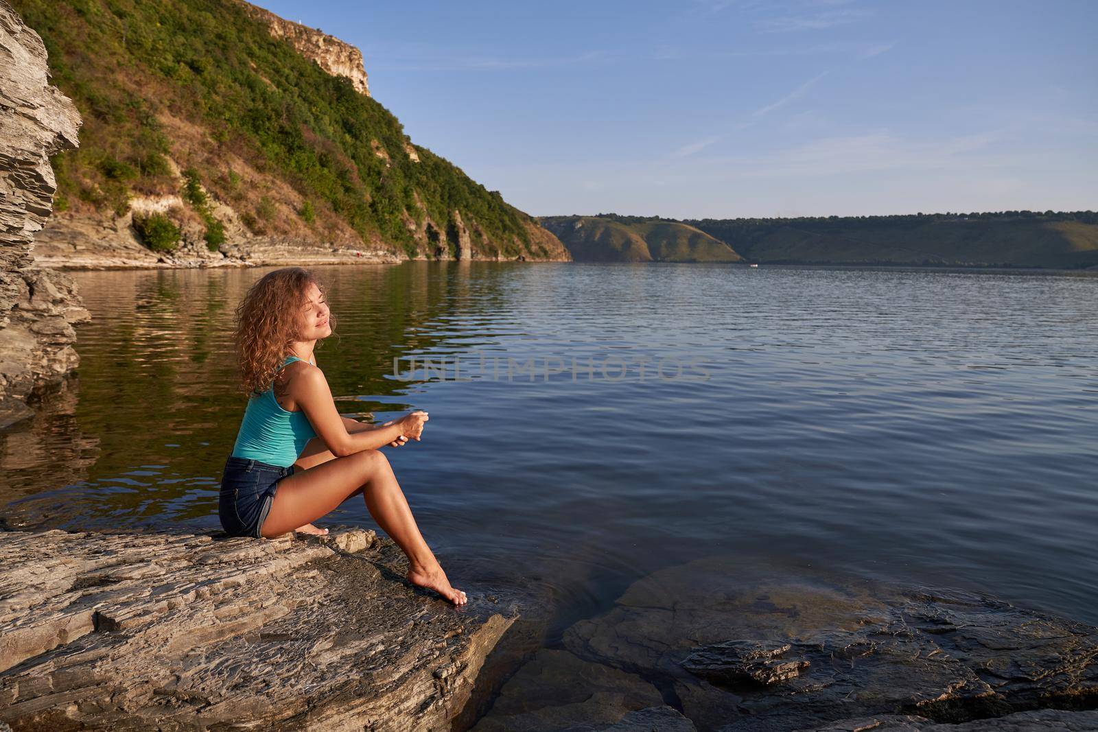 Romantic, pretty girl with long curly hair sitting bare foot on shore of lake. Beautiful, seductive model wearing in blue shorts and t shirt resting, admiring landscape.