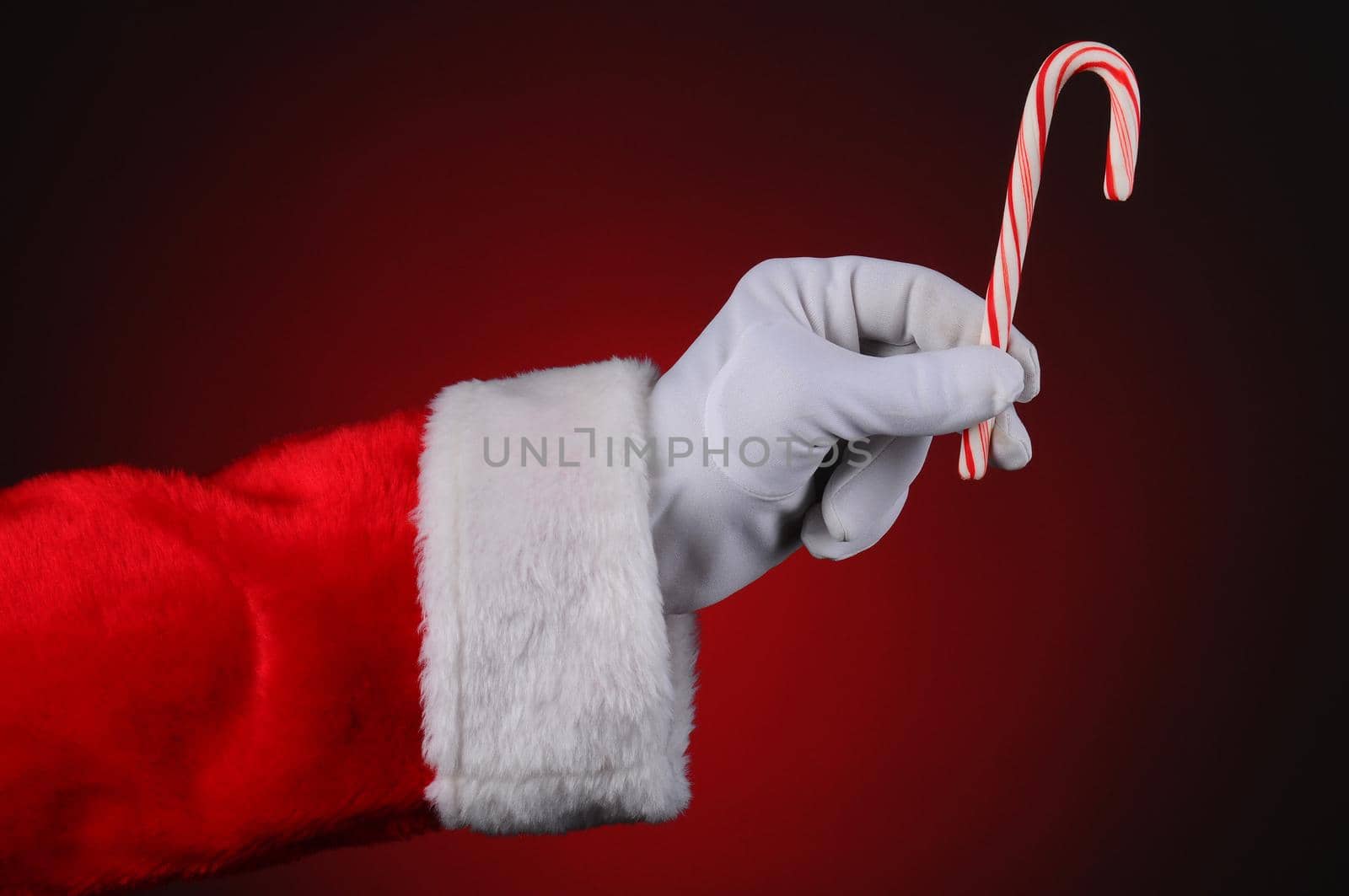 Santa Claus hand holding a candy cane over a red light to dark background. Horizontal format showing hand and arm only.