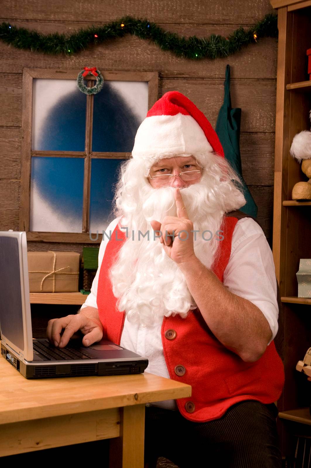 Santa Claus in Workshop Using Laptop and making Shh sign at viewer. Vertical Composition