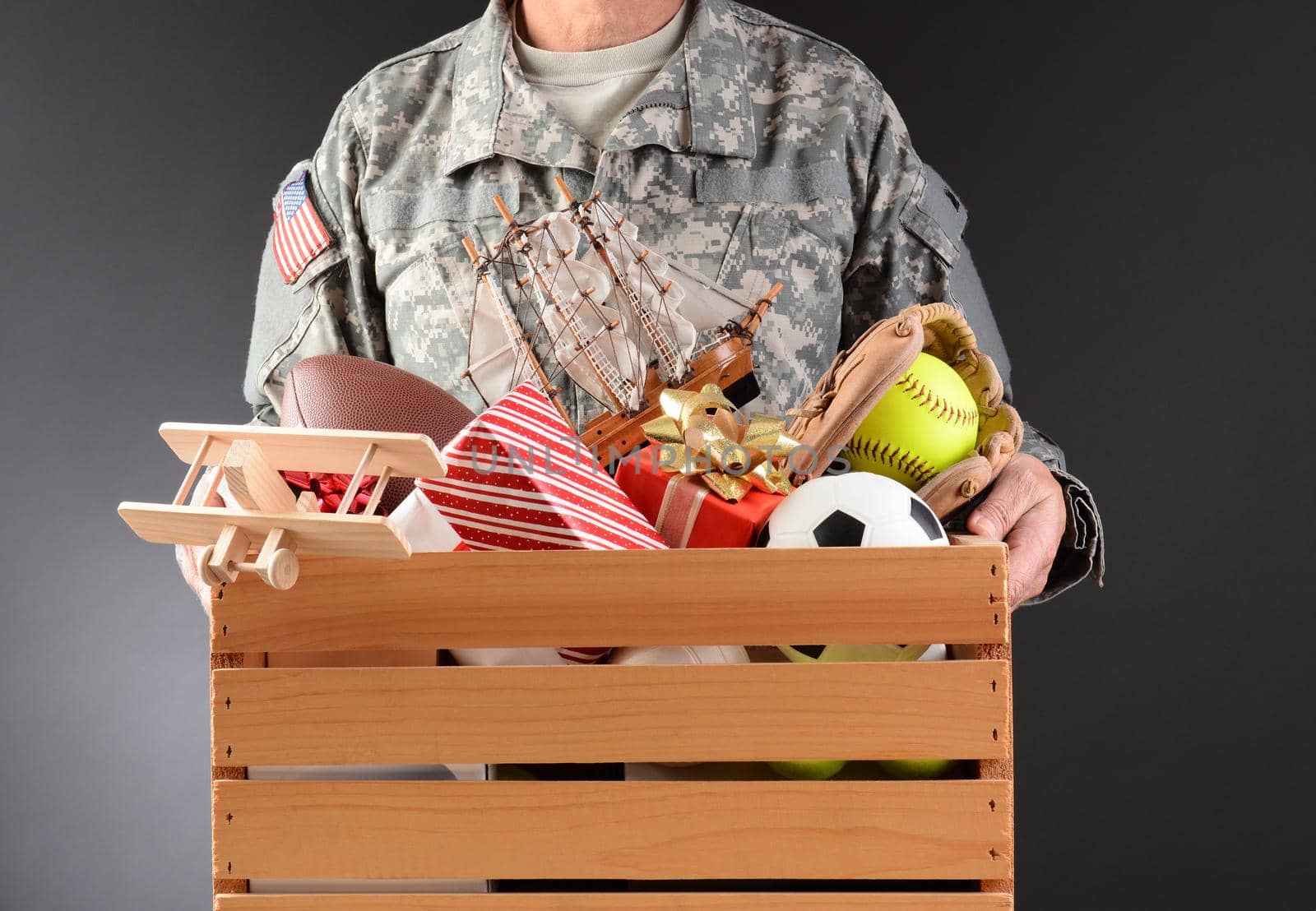 Closeup of a soldier in fatigues holding a wooden box full of toys and sports equipment for a holiday charity drive. Horizontal format man is unrecognizable. 