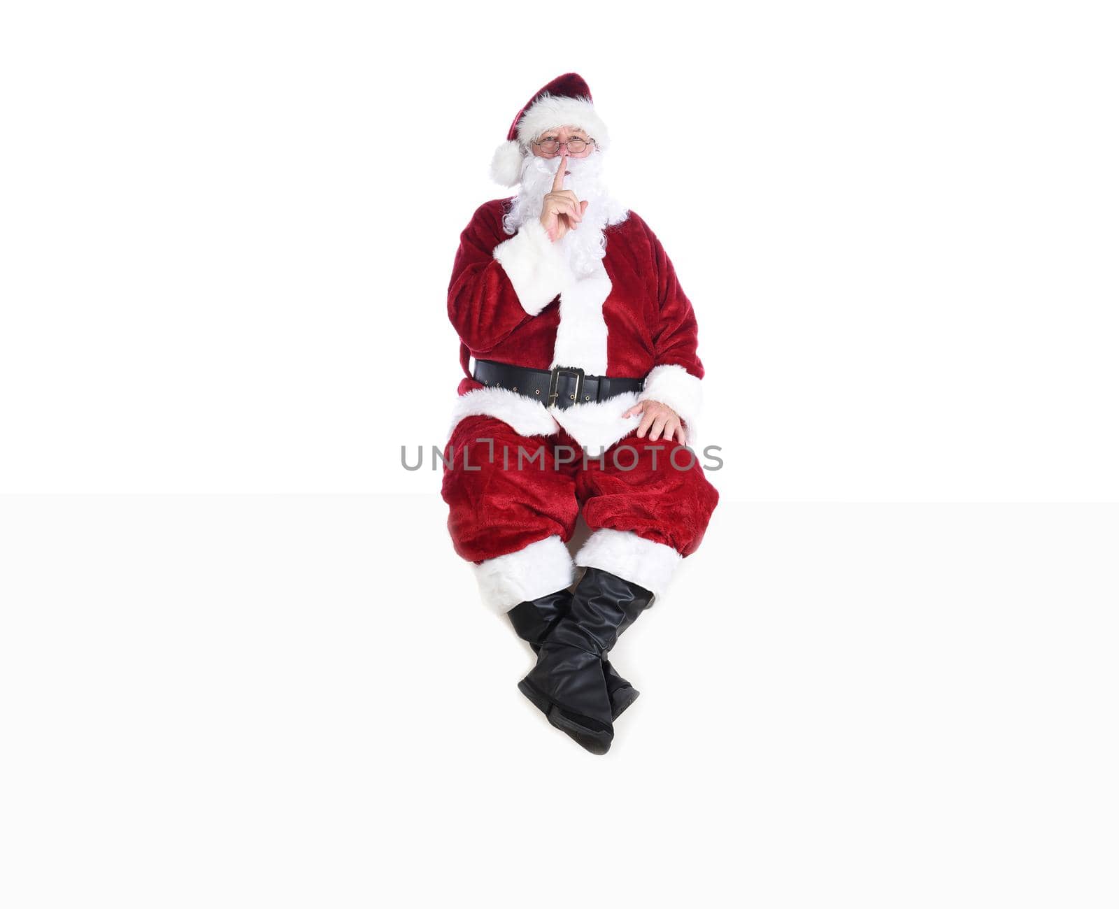 Senior man in traditional Santa Claus Suit sitting on a white wall with a finger to his lips making a Shh Gesture. by sCukrov