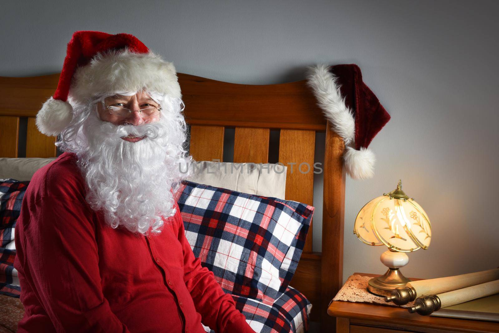 Closeup of Santa Claus wearing a red union suit sitting on the edge of his bed at the North Pole, ready to go to sleep after delivering packages on Christmas eve. by sCukrov