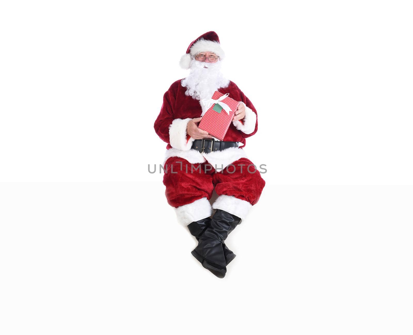 Senior man in traditional Santa Claus Suit sitting on a white wall and holding a wrapped present. by sCukrov