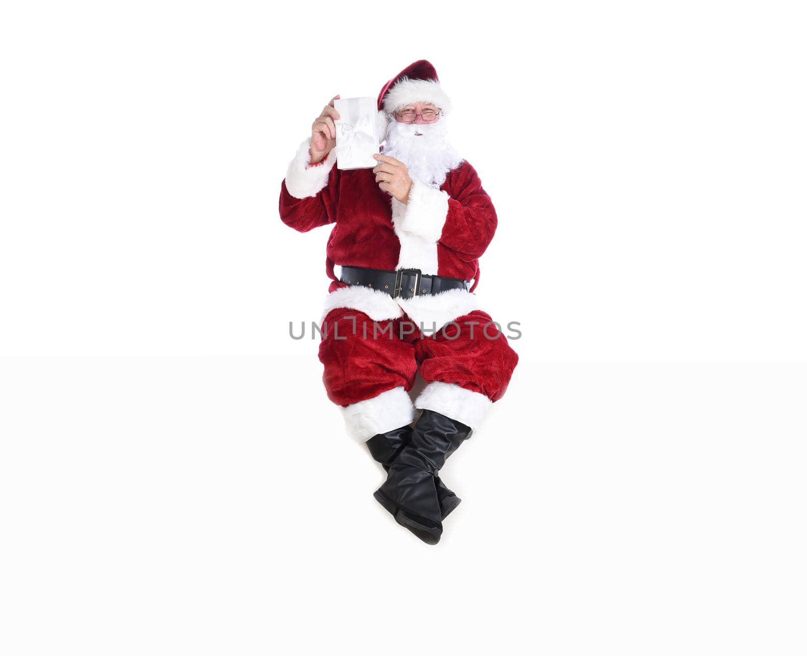 Senior man in traditional Santa Claus Suit sitting on a white wall holding a wrapped Christmas Present.  Isolated on white with copy space. by sCukrov