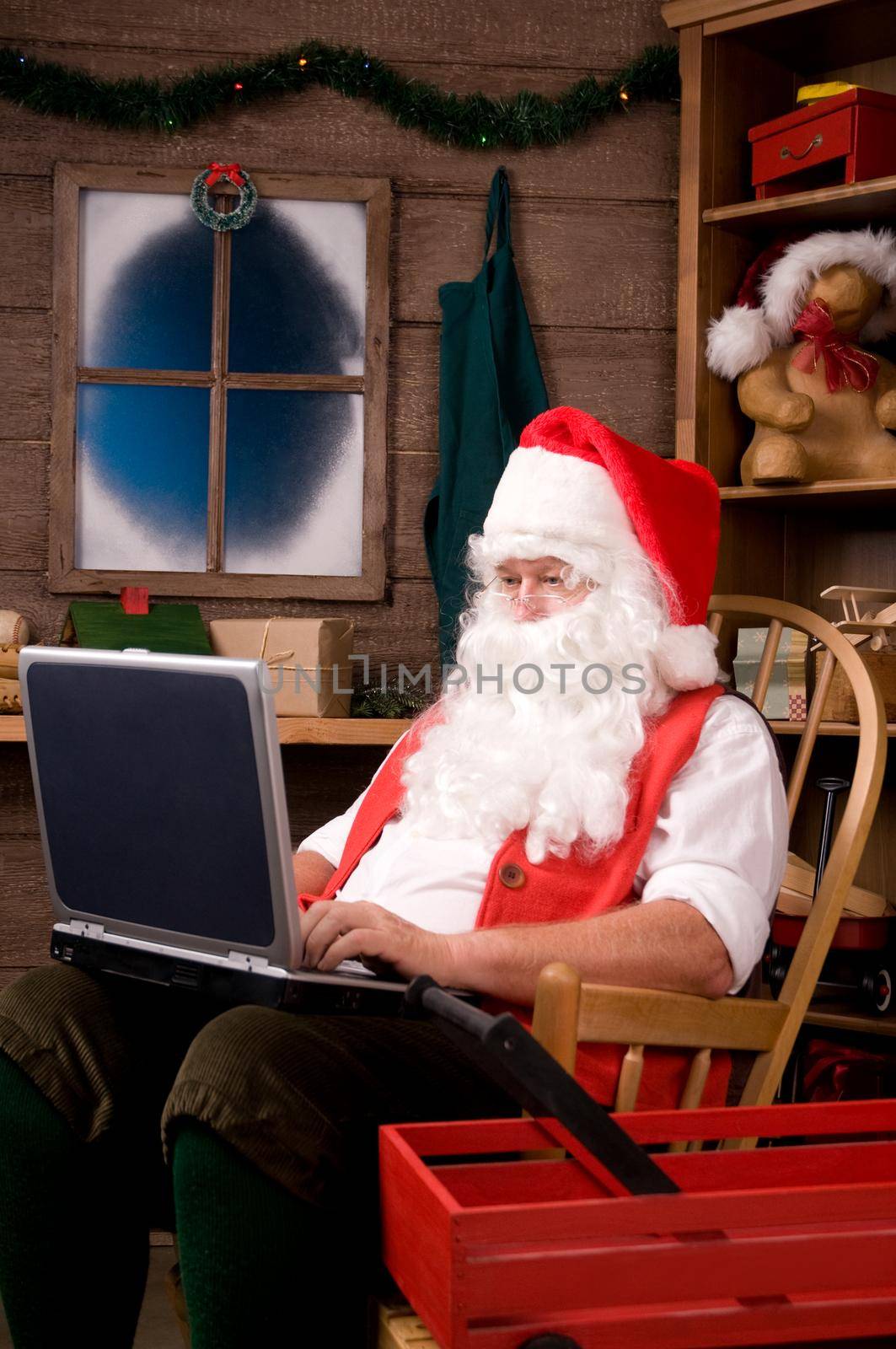 Santa Claus Sitting in Rocking Chair in Workshop Using Laptop. Vertical composition.