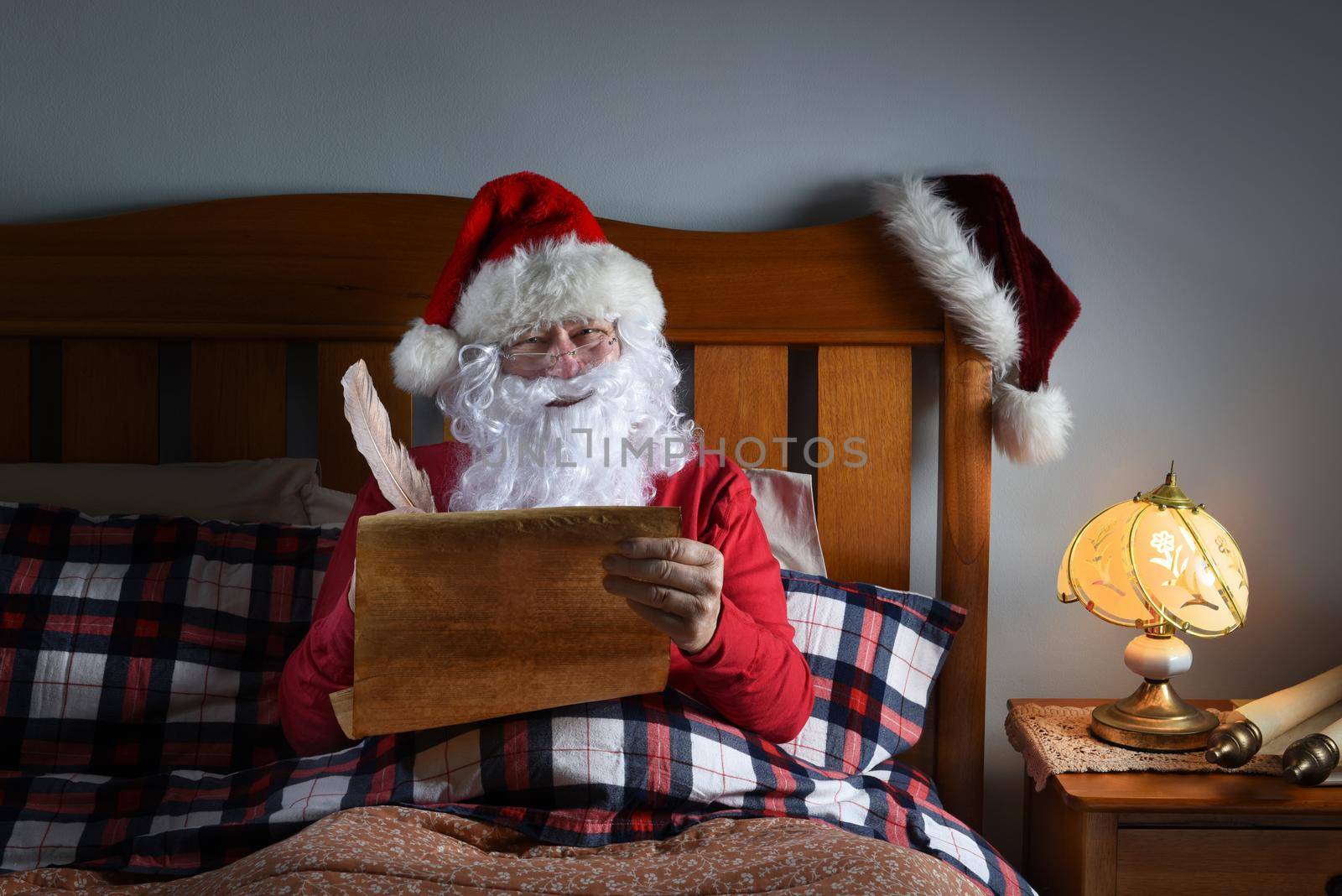 Santa Claus sitting up in his bed working on his naughty and nice list. by sCukrov