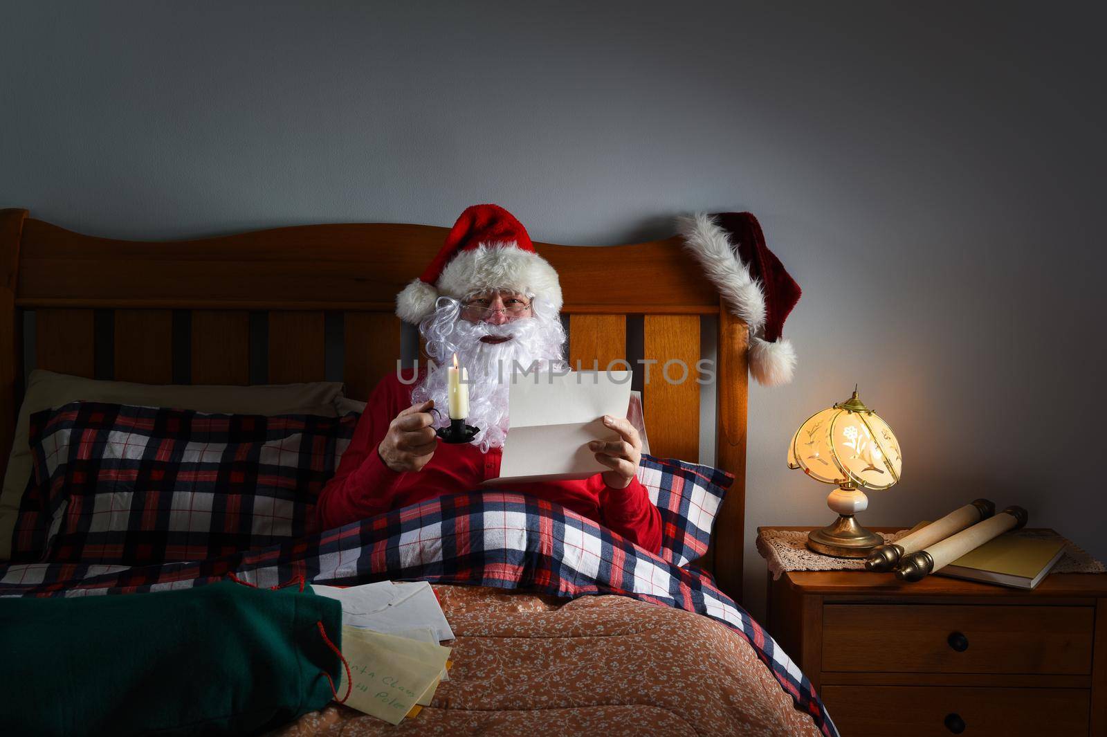 Santa Claus reading letters by candlelight in bed at his North Pole home.