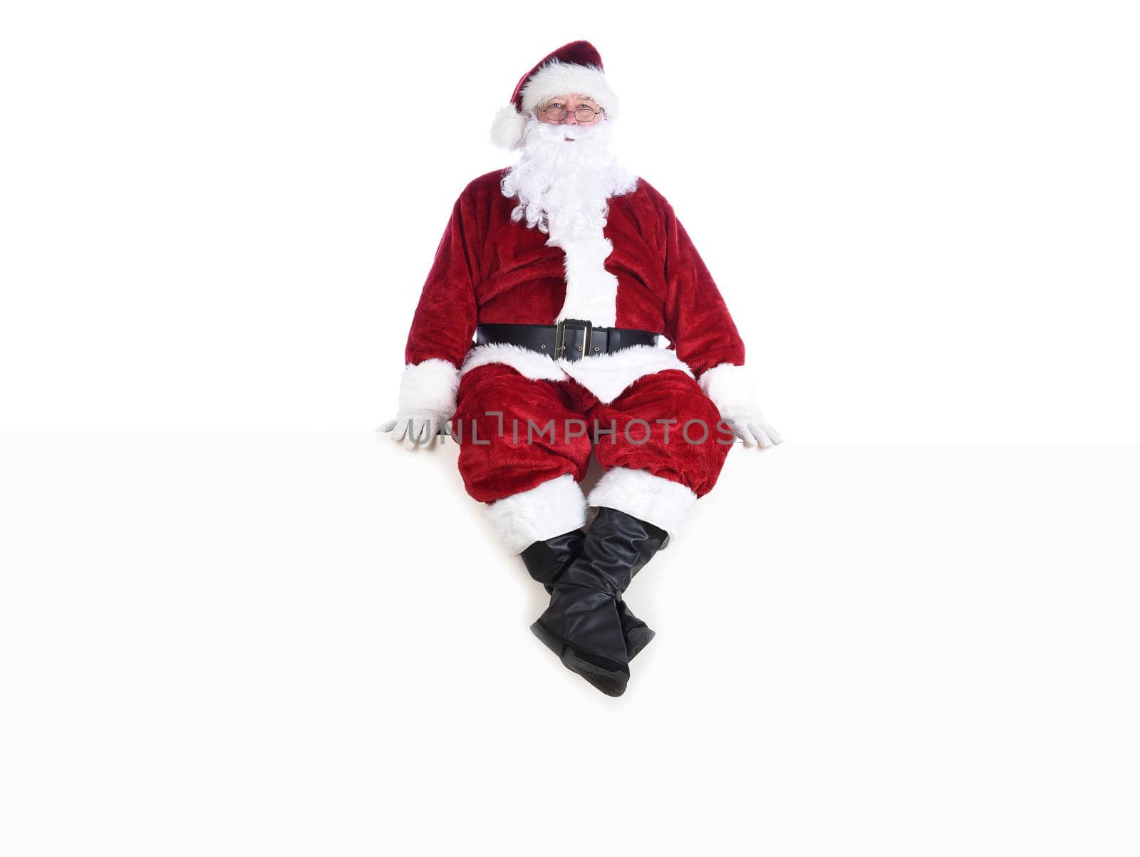 Senior man in traditional Santa Claus Suit sitting on a white wall.  Isolated on white with copy space.