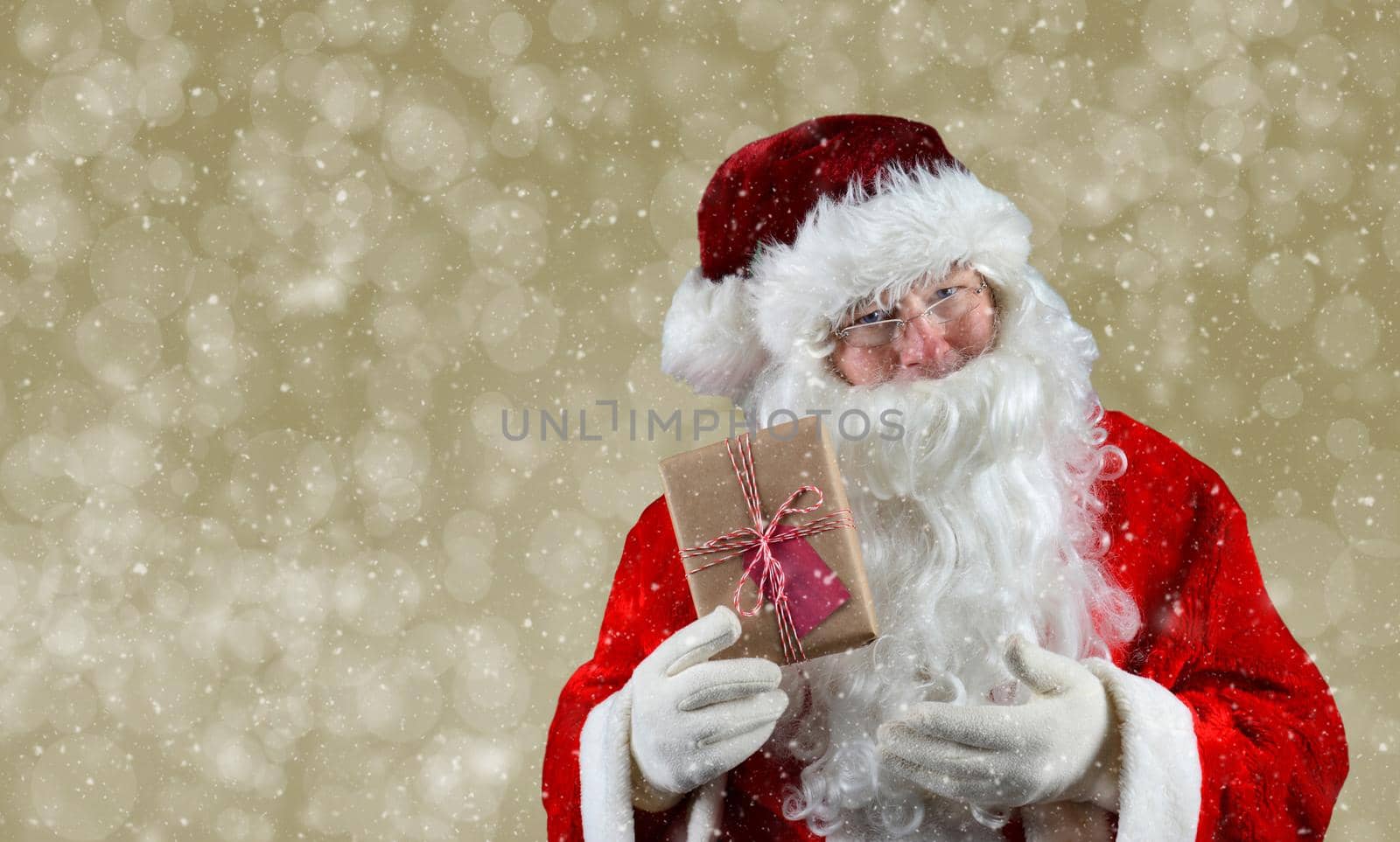 Santa Claus holding a plain brown wrapped package. The eco friendly recyclable gift is tied with string and has a blank gift tag. On a light gold bokeh background.