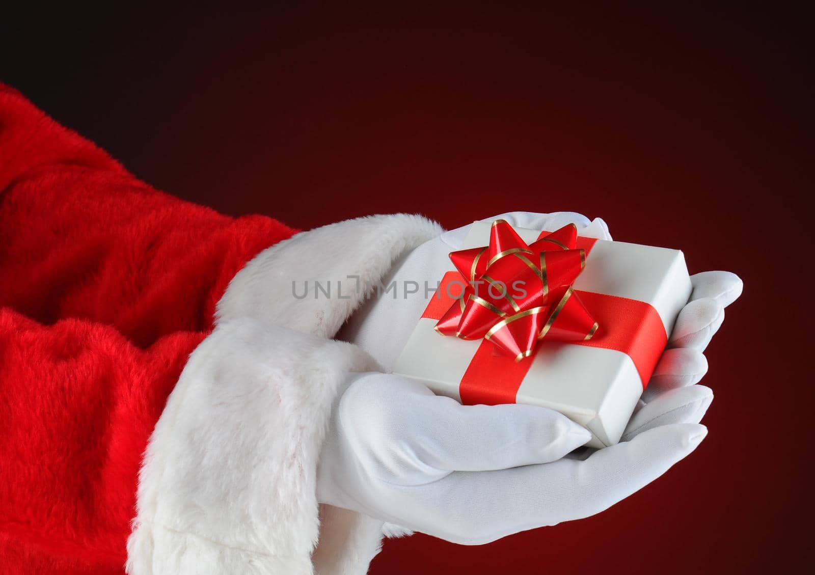 Santa Claus holding a small Christmas Present in both of his hands. Horizontal format showing only hand and arms on a light ot dark red background.