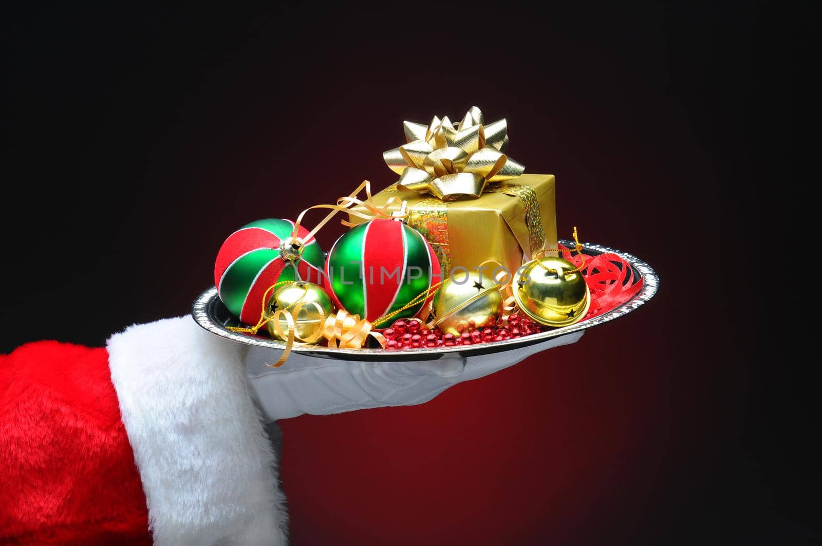 Santa Claus hand holding a tray with decorations and a gift. Hand and arm only on a light to dark red background.