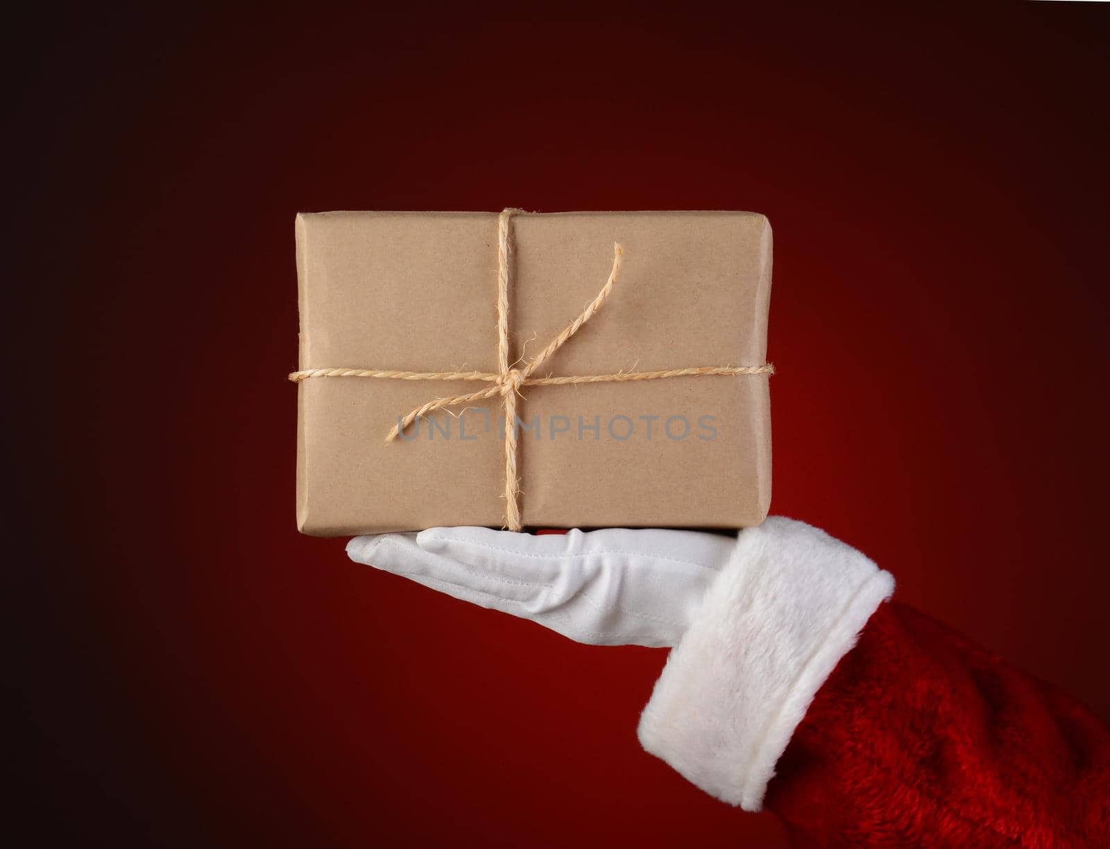 Closeup of Santa Claus holding a parcel in the palm of his outstretched hand. Hand and arm only over a light to dark red spot background.