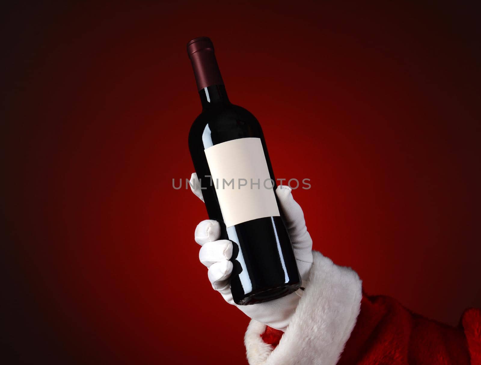 Closeup of Santa Claus holding a bottle of wine in his hand. Hand and arm only over a light to dark red spot background.