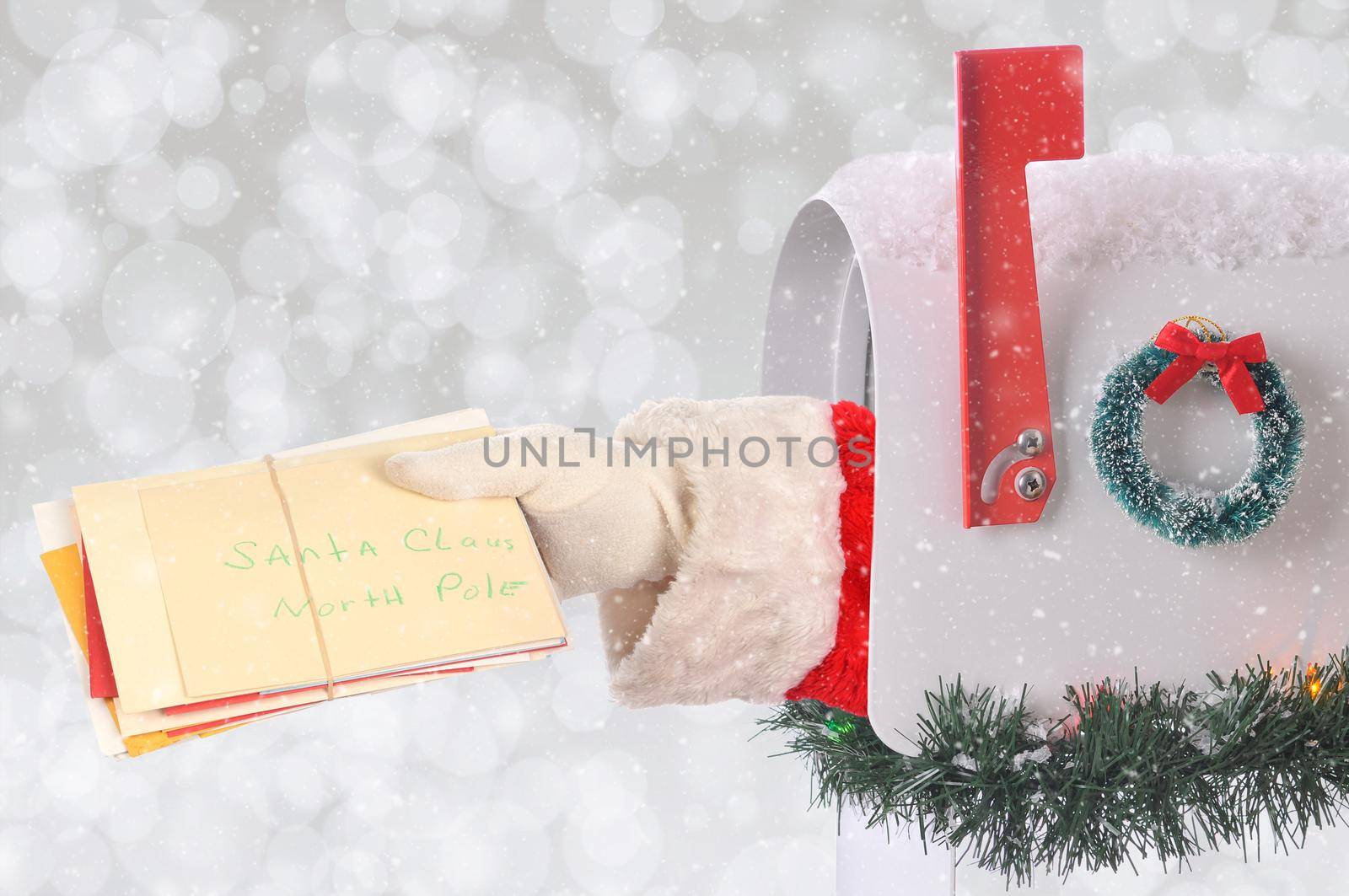 Santa Claus arm holding a stack of Christmas letters coming out of a mail box with silver bokeh background and snow effect. by sCukrov