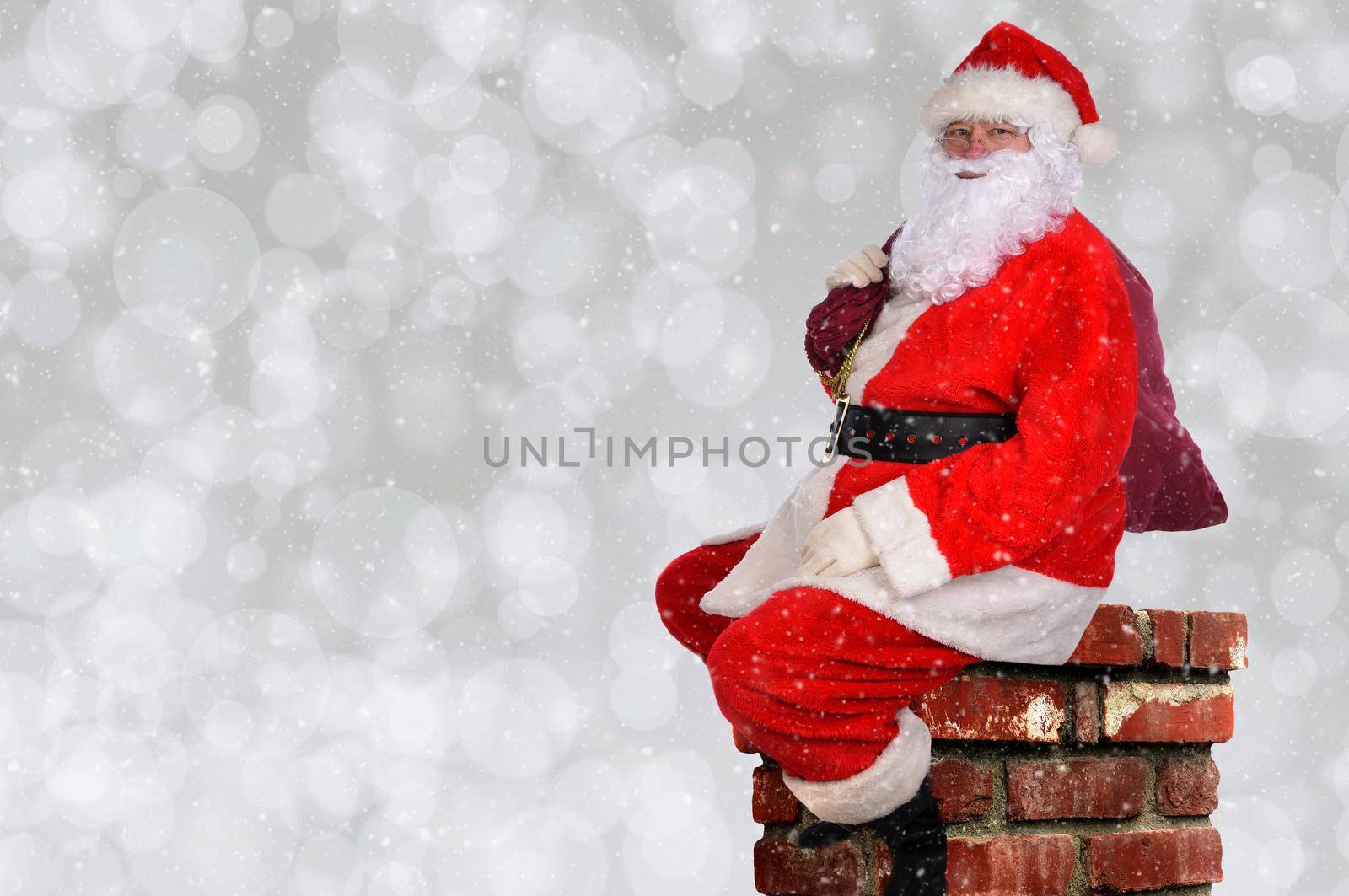 Santa Claus with his bag of toys sitting atop a chminey, with silver bokeh background and snow flake effect.