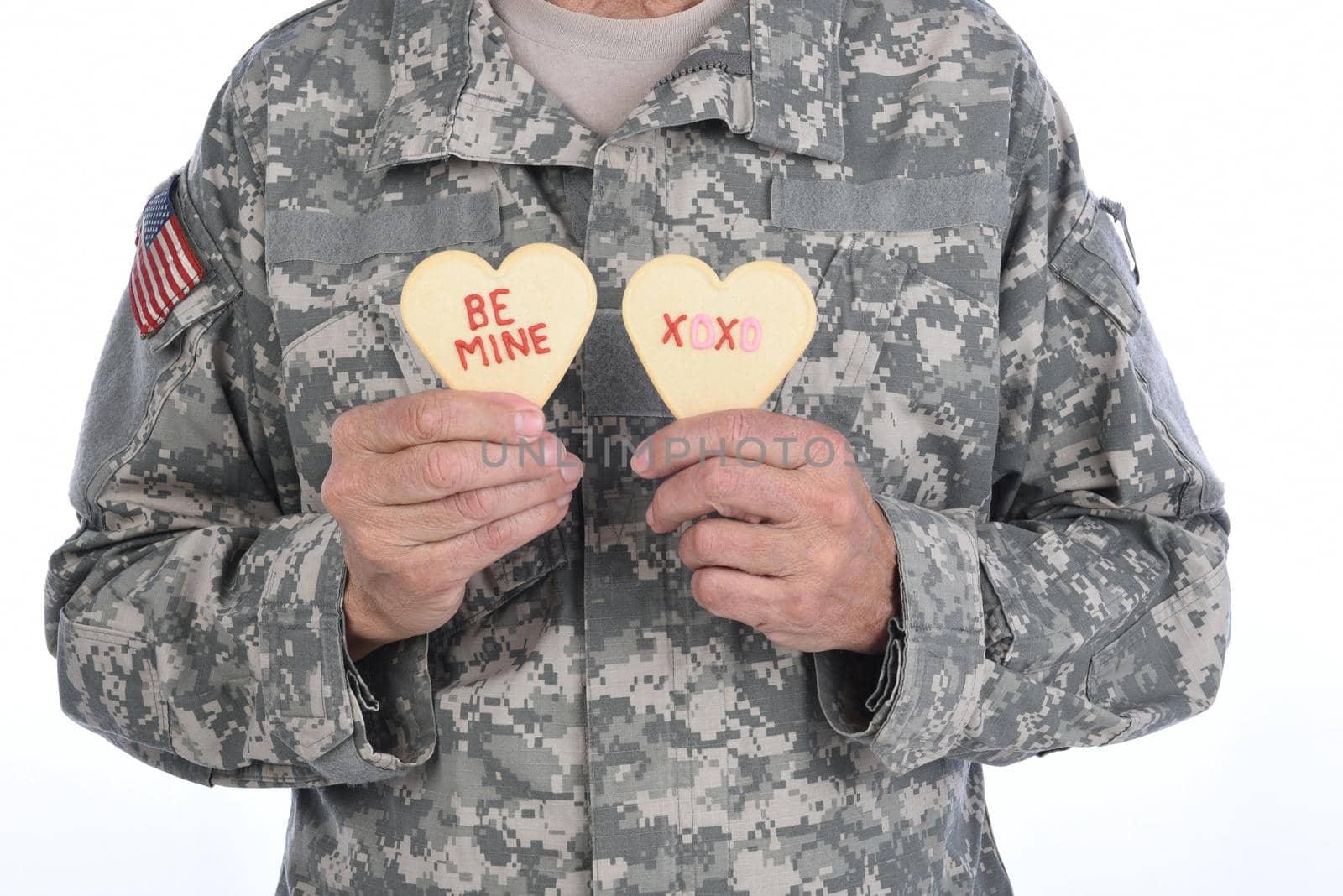 Closeup of a soldier holding two Heart Shaped Valentines Day cookies with the words Be Mine and XOXO written in red icing.