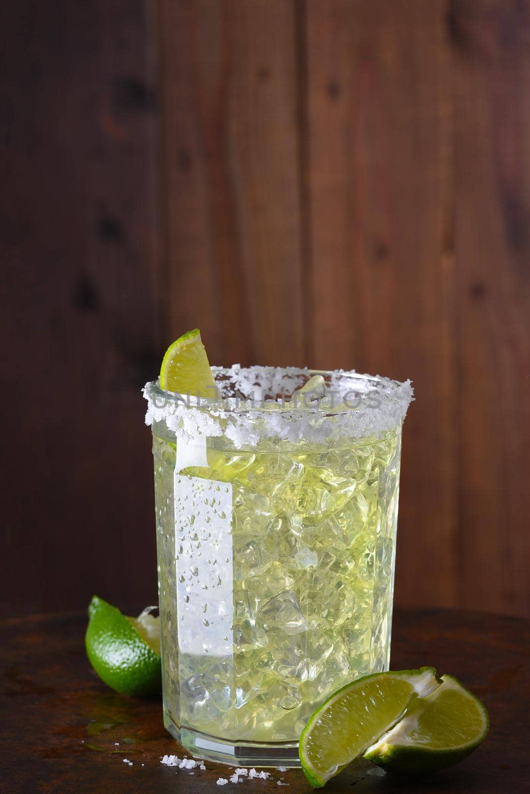 A margarita on the rocks in a glass tumbler. The rim of the glass is encrusted in salt and sliced limes are on the bar surface.