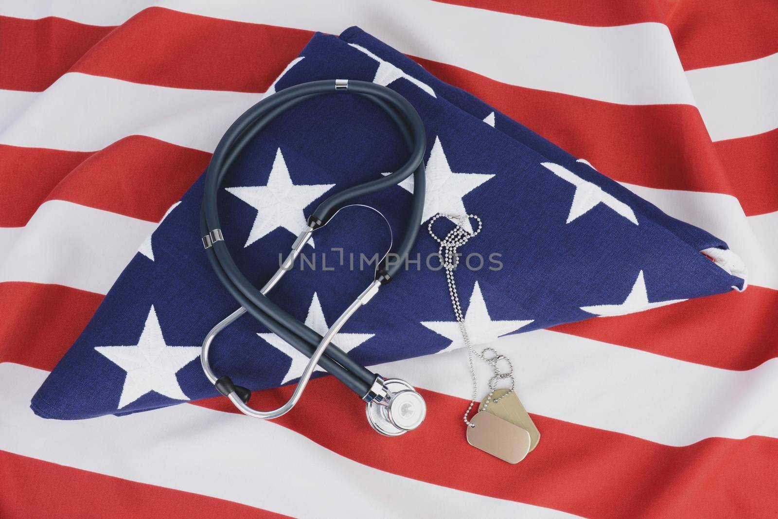 A stethoscope and dog tags on a folded American Flag. The background is the red and white stripes of another flag. Ideal for Military Health care concepts.