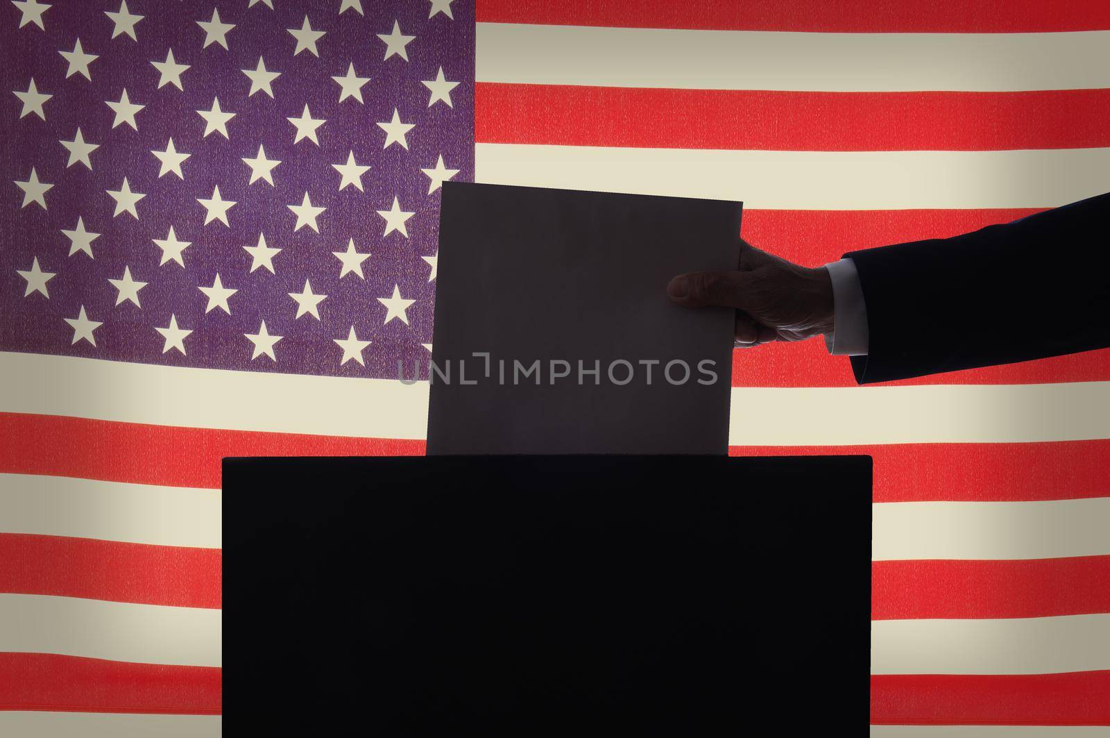 Voting Concept. Closeup of a man’s hand, in silhouette,  placing a ballot into a box in front of an American Flag.  by sCukrov