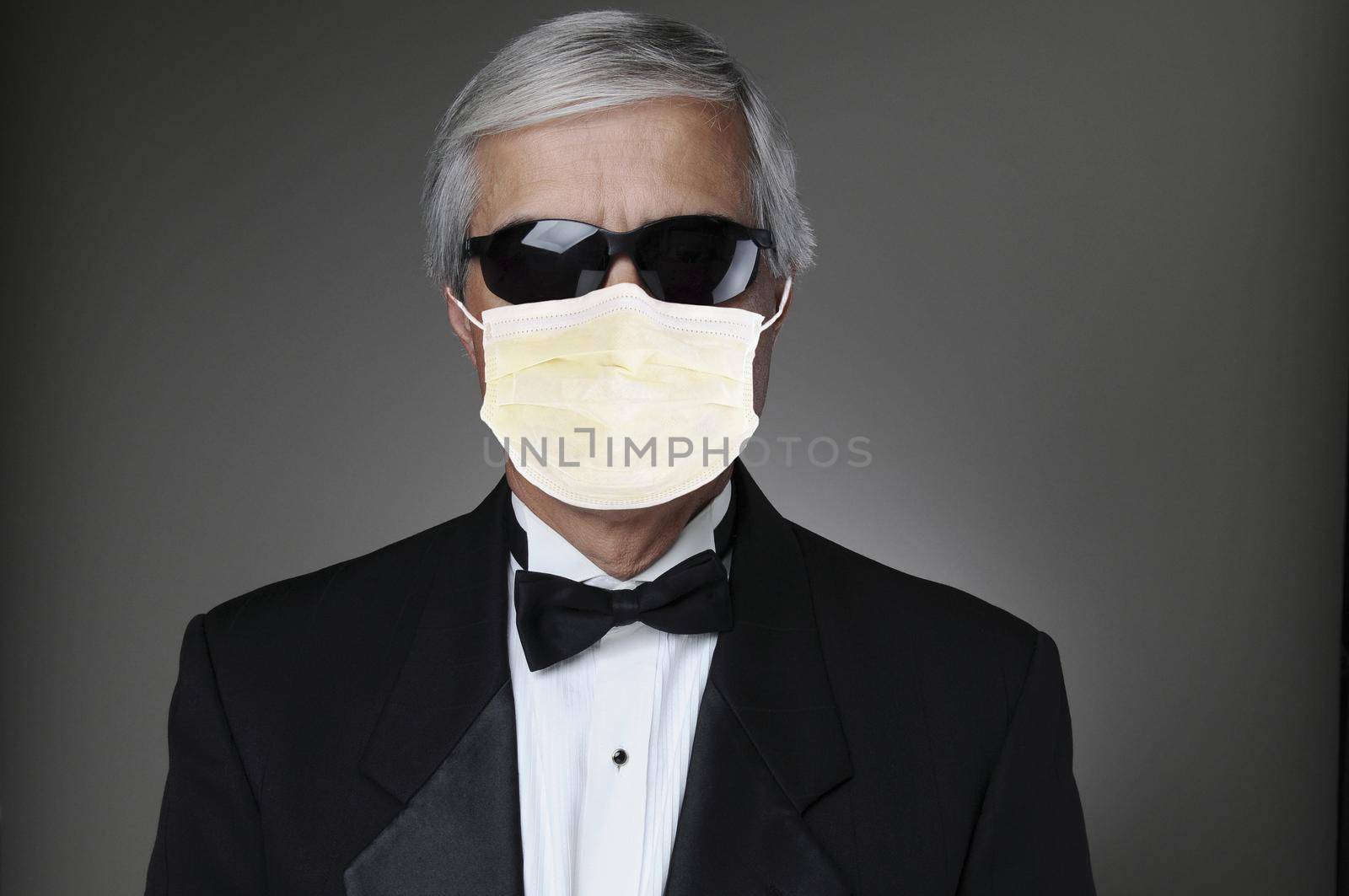 Portrait of a middle aged man in a tuxedo wearing a COVID-19 protective mask and sunglasses. Horizontal format over a gray background.