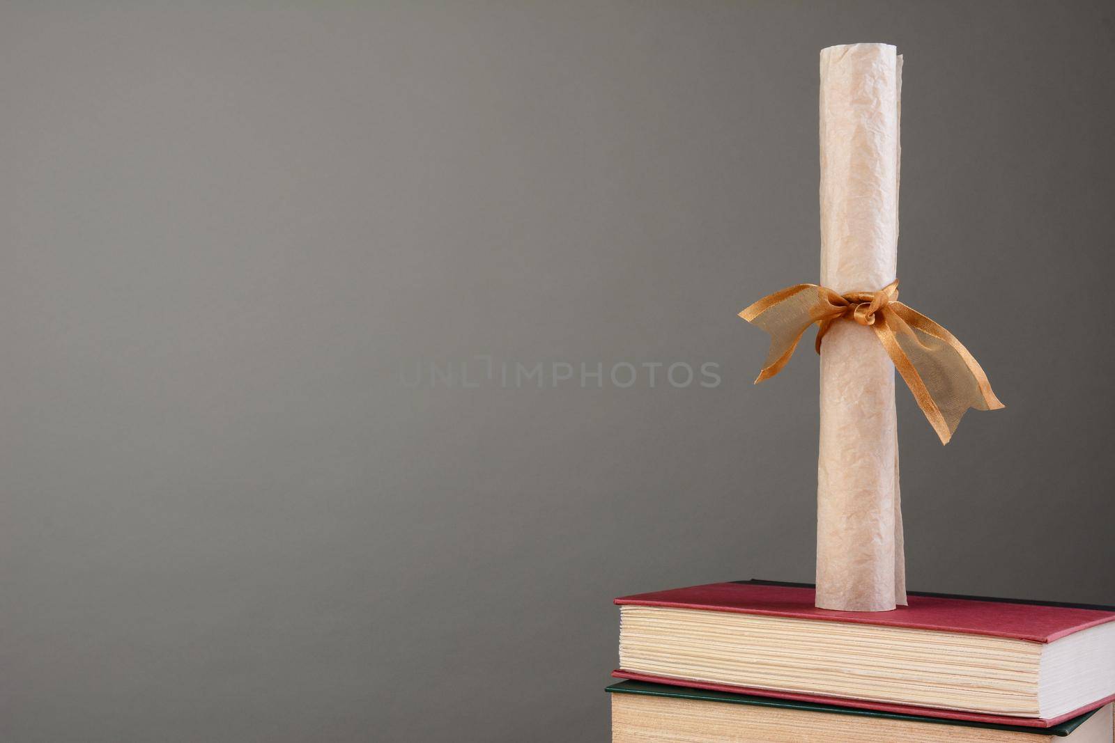 A parchment diploma tied with a gold ribbon standing on a stack of school books against a light to dark gray background. Horizontal format with copy space.
