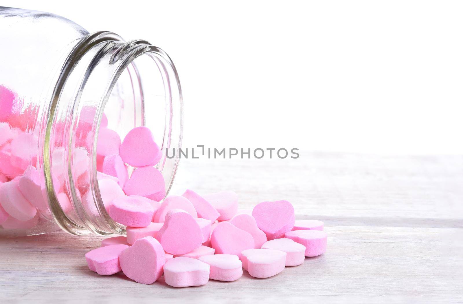 Closeup of Pink Valentine's Day pink candy hearts spilling out of a jar onto a rustic white wood table with copy space.