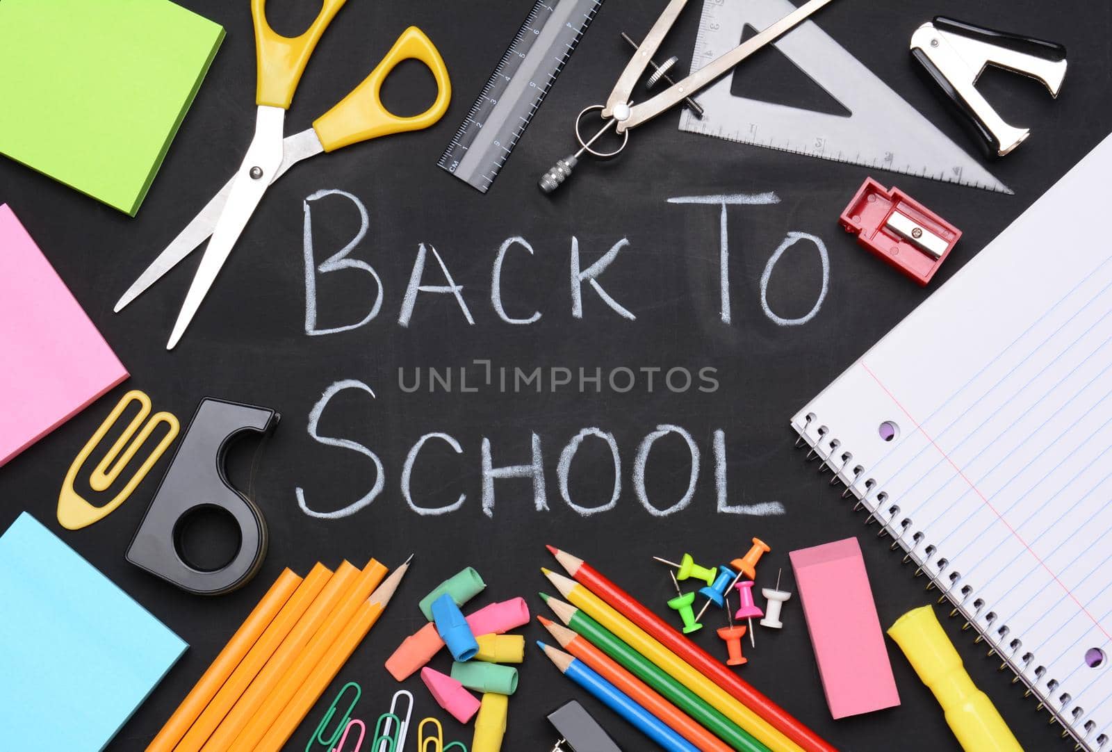 Back to school written on a chalkboard. The words are surrounded by school supplies including, paper, scissors, pencils, erasers, paper clips, compass, ruler, push pins and other necessities for students.