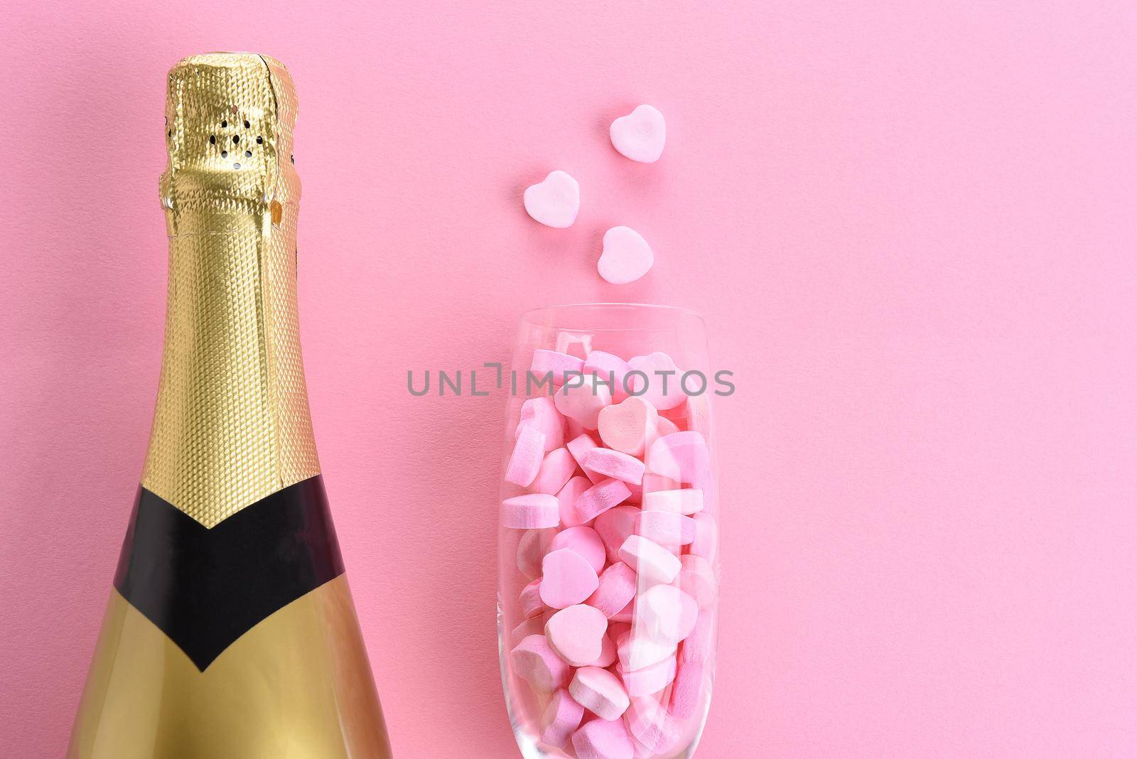 Closeup of a bottle of champagne and a flute filled with pink candy hearts for Valentines Day. On a pink background with copy space.