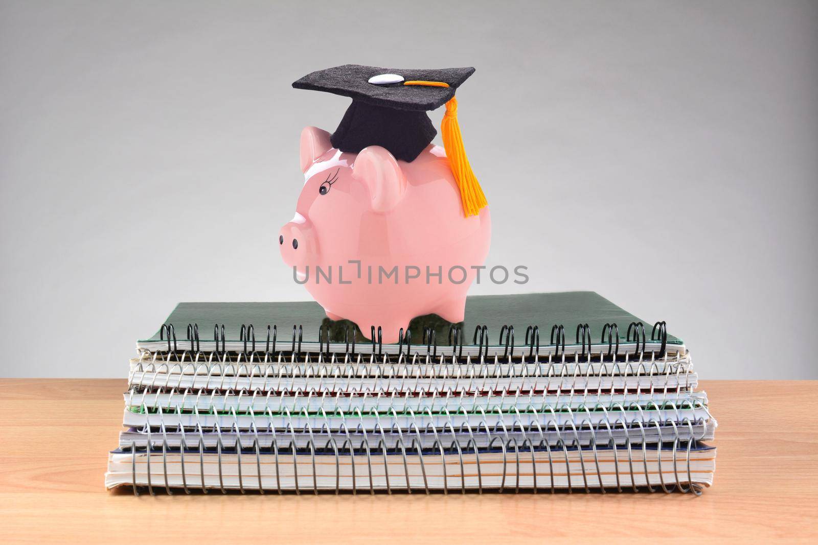 Closeup of a stack of spiral notebooks on a teachers desk with a piggy bank wearing a graduation cap, representing the high cost of education. by sCukrov