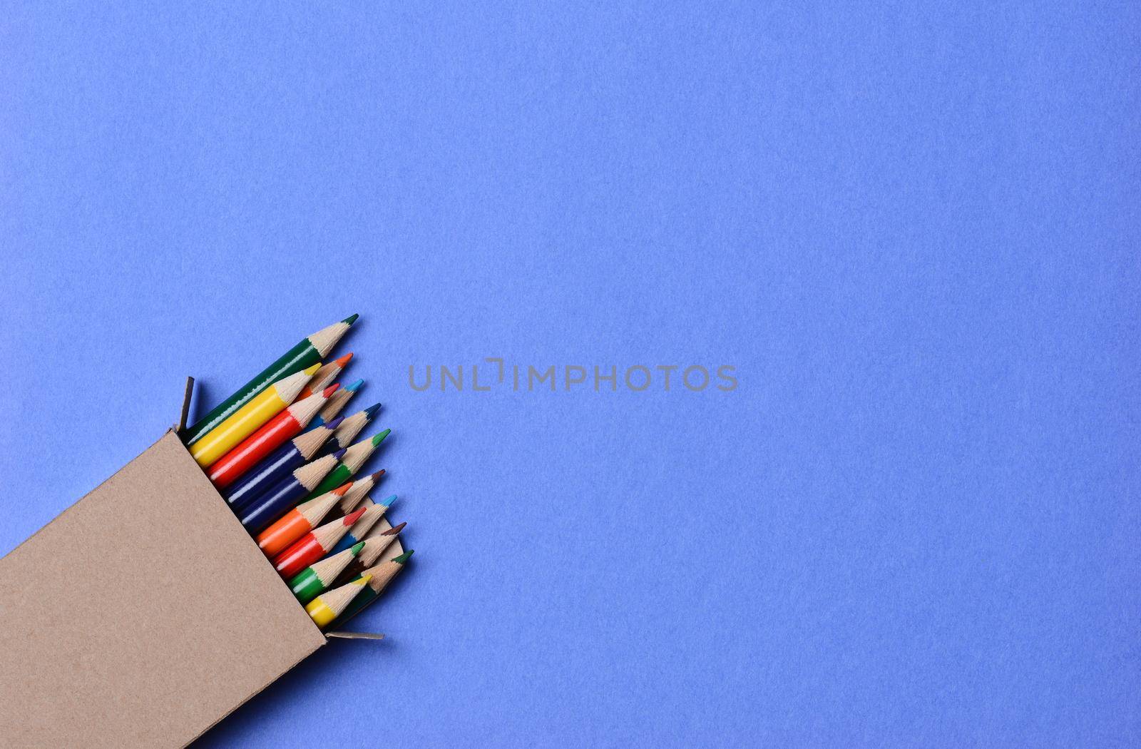 High angle shot of a box of multi-colored pencils on a blue background. The pencils are part way out of the box which is at an angle in the lower left corner leaving copy space.