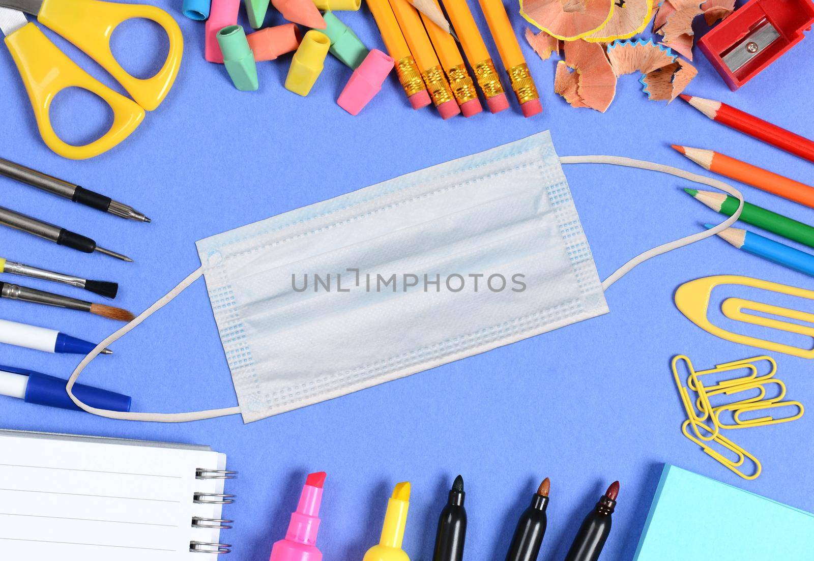 COVID-19 Mask with Back to School supplies arranged on the outer edge of a blue background. 