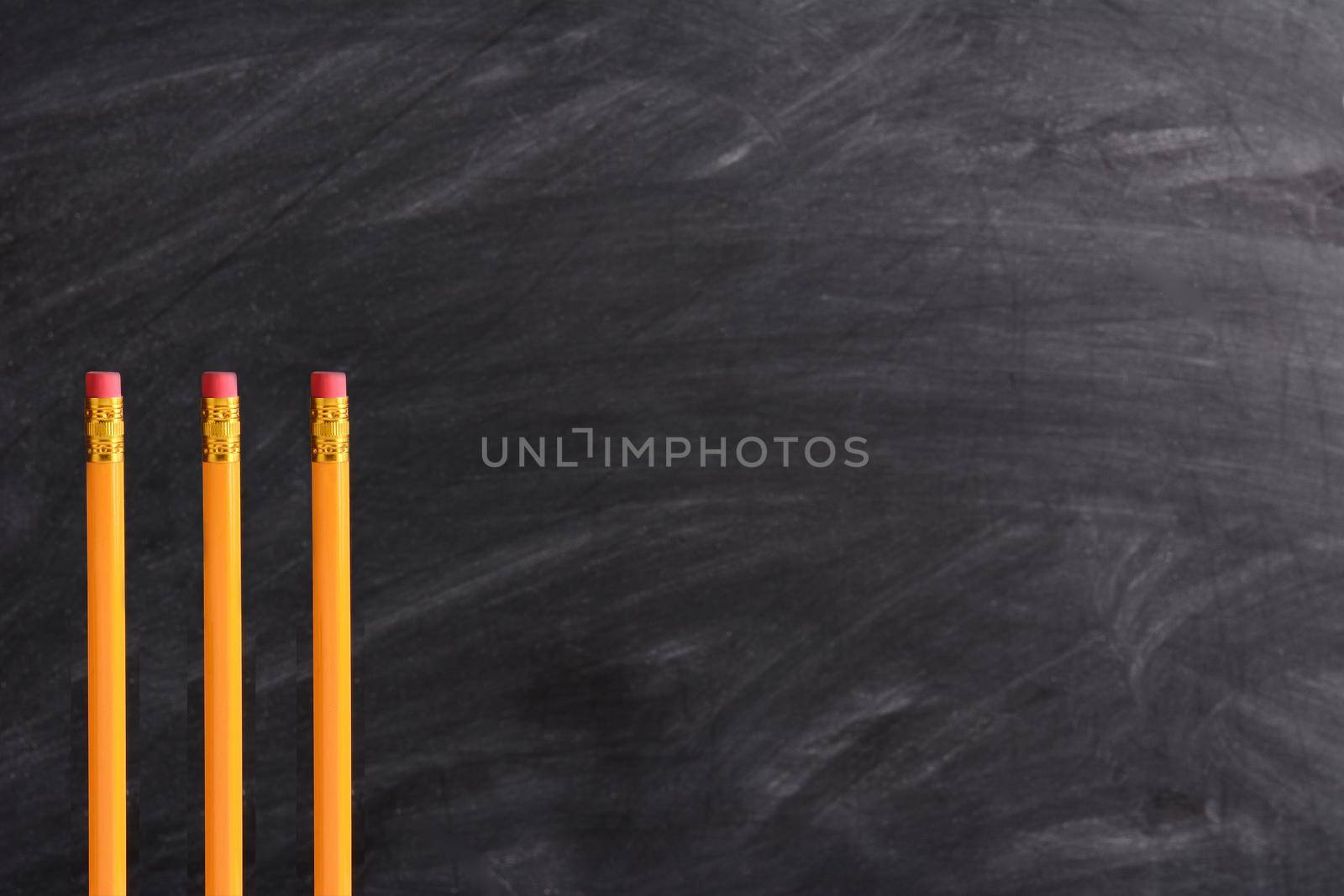 Three new pencils standing in front of a chalkboard with eraser marks. Horizontal format with copy space. Back to School concept.