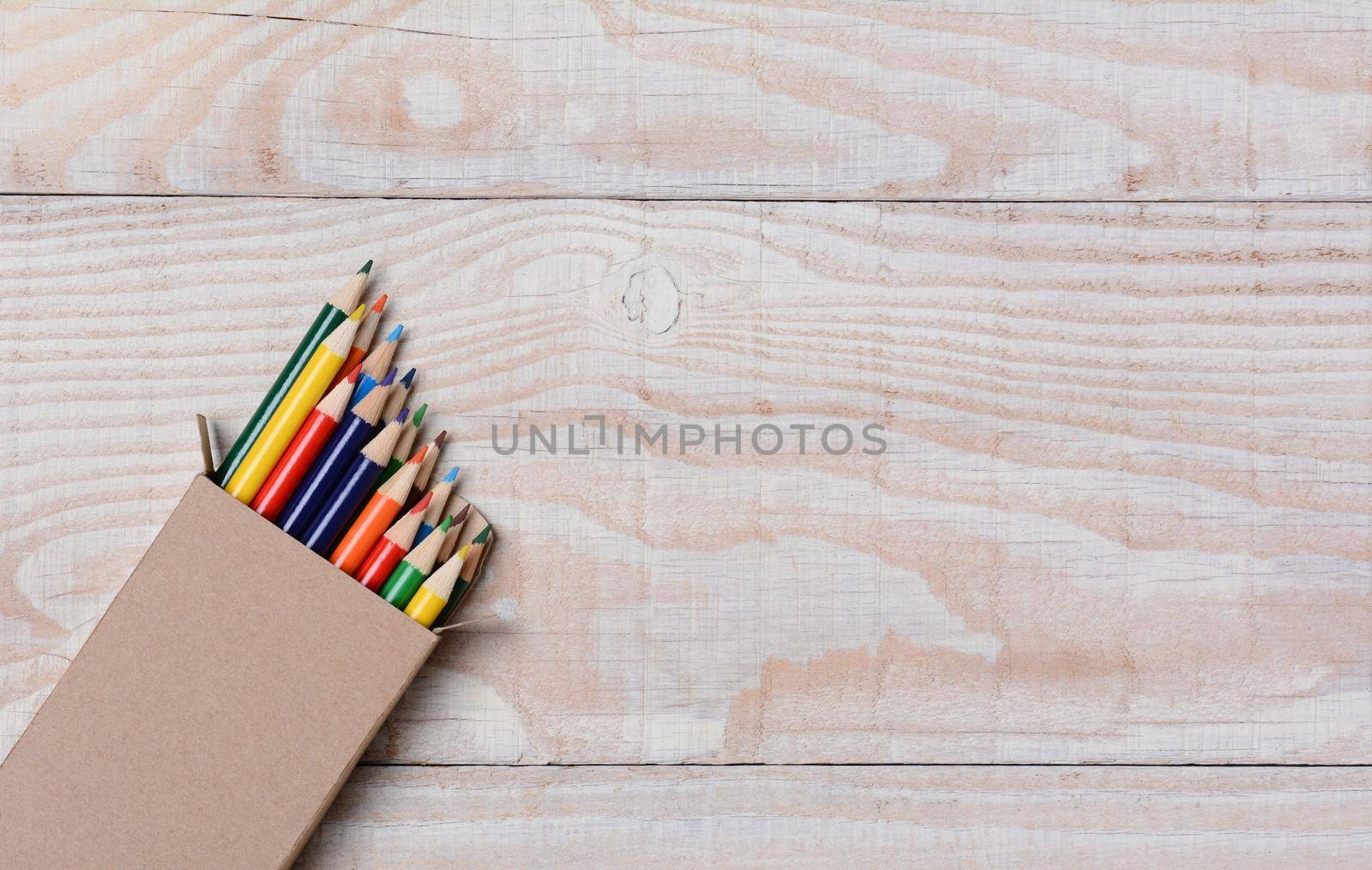 High angle shot of a box of multi-colored pencils on a white wood table. The pencils are partially out of the box and at an angle in the bottom left corner, with copy space.