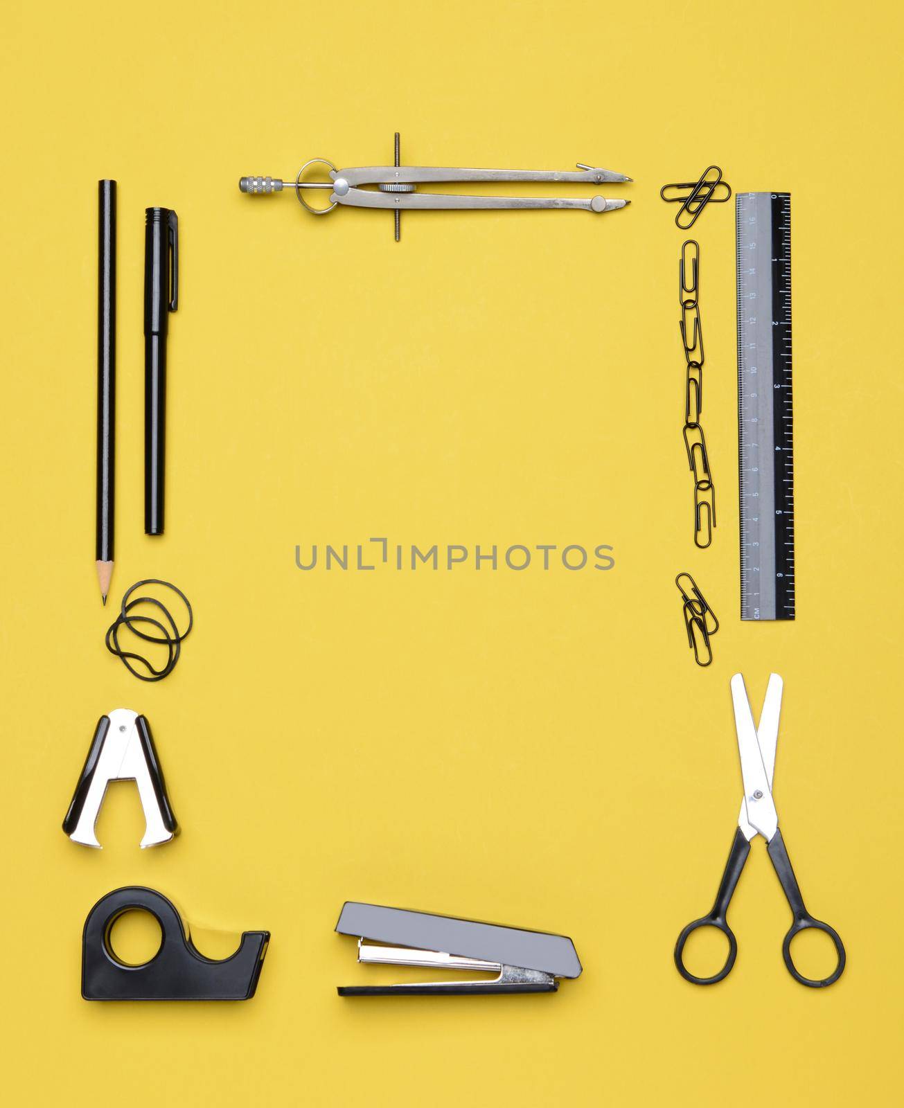 Office and back to school supplies on a yellow background. Looking down on the all black and chrome tools from an overhead angle. The items are arranged in a rectangle forming a frame.
