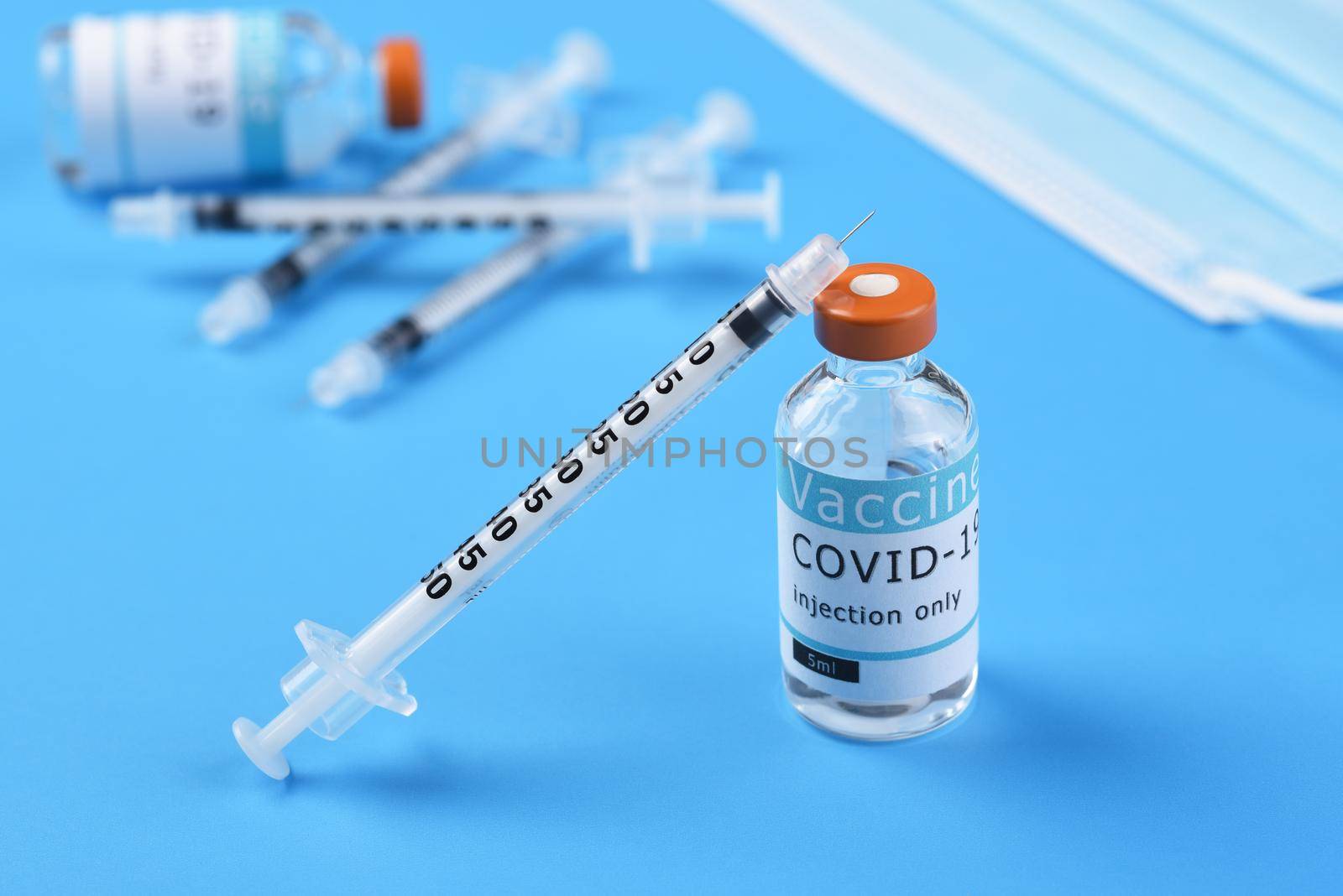 A vial of Covid-19 vaccine with a syringe leaning on the bottle with more syringes, a bottle and surgical mask in the background.  by sCukrov
